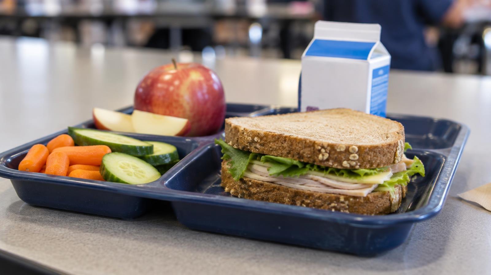 School lunch tray with a sandwich, sliced fruit, vegetables, and a milk carton on a cafeteria table.