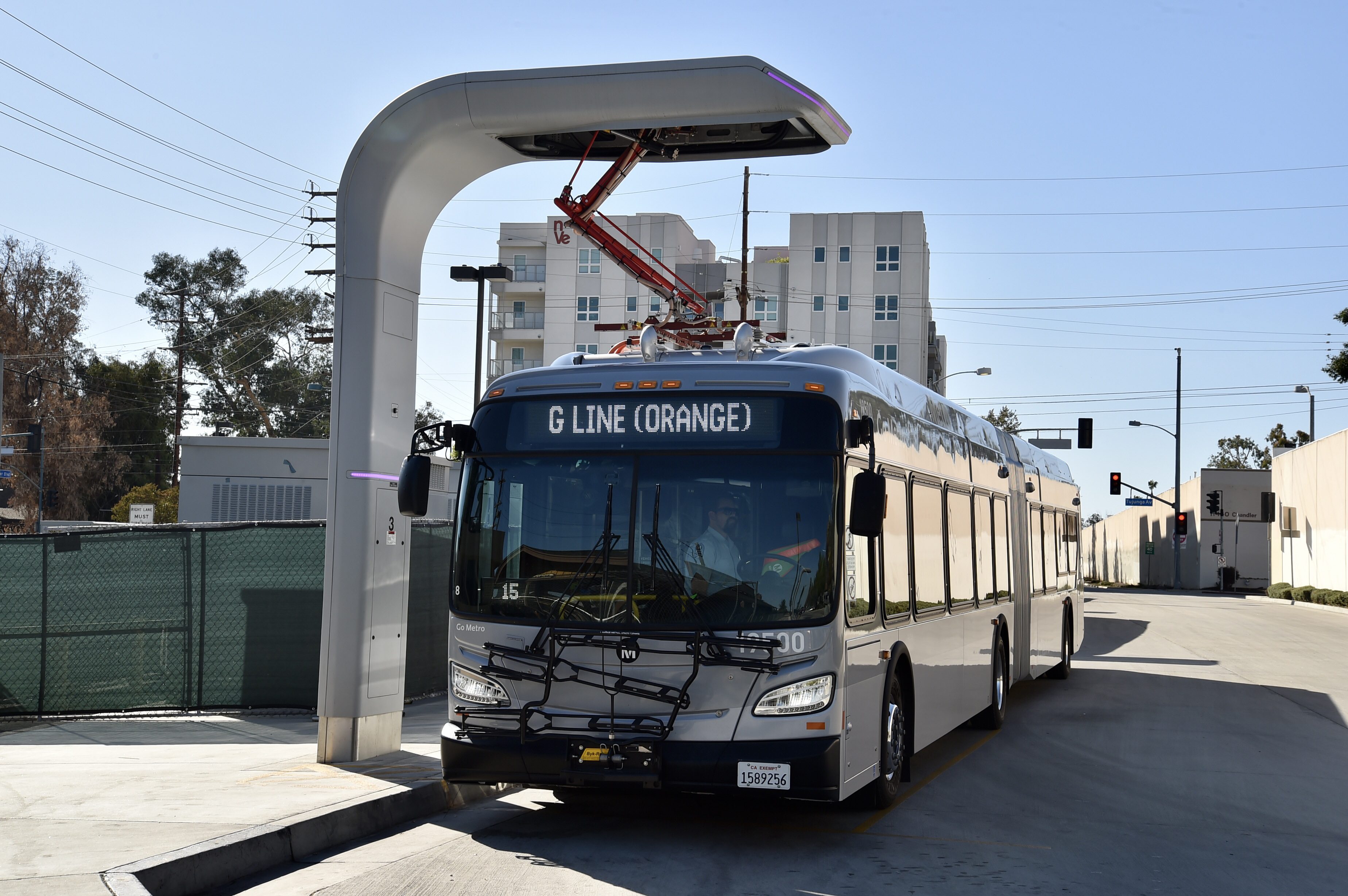 An electric articulated bus connected to an overhead charging arch at a transit station.