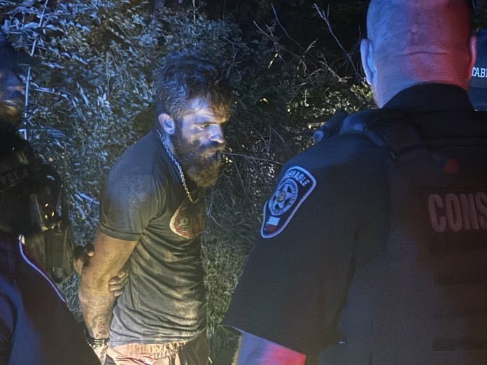 Handcuffed man in gray shirt detained by uniformed constable officers at night outdoors.