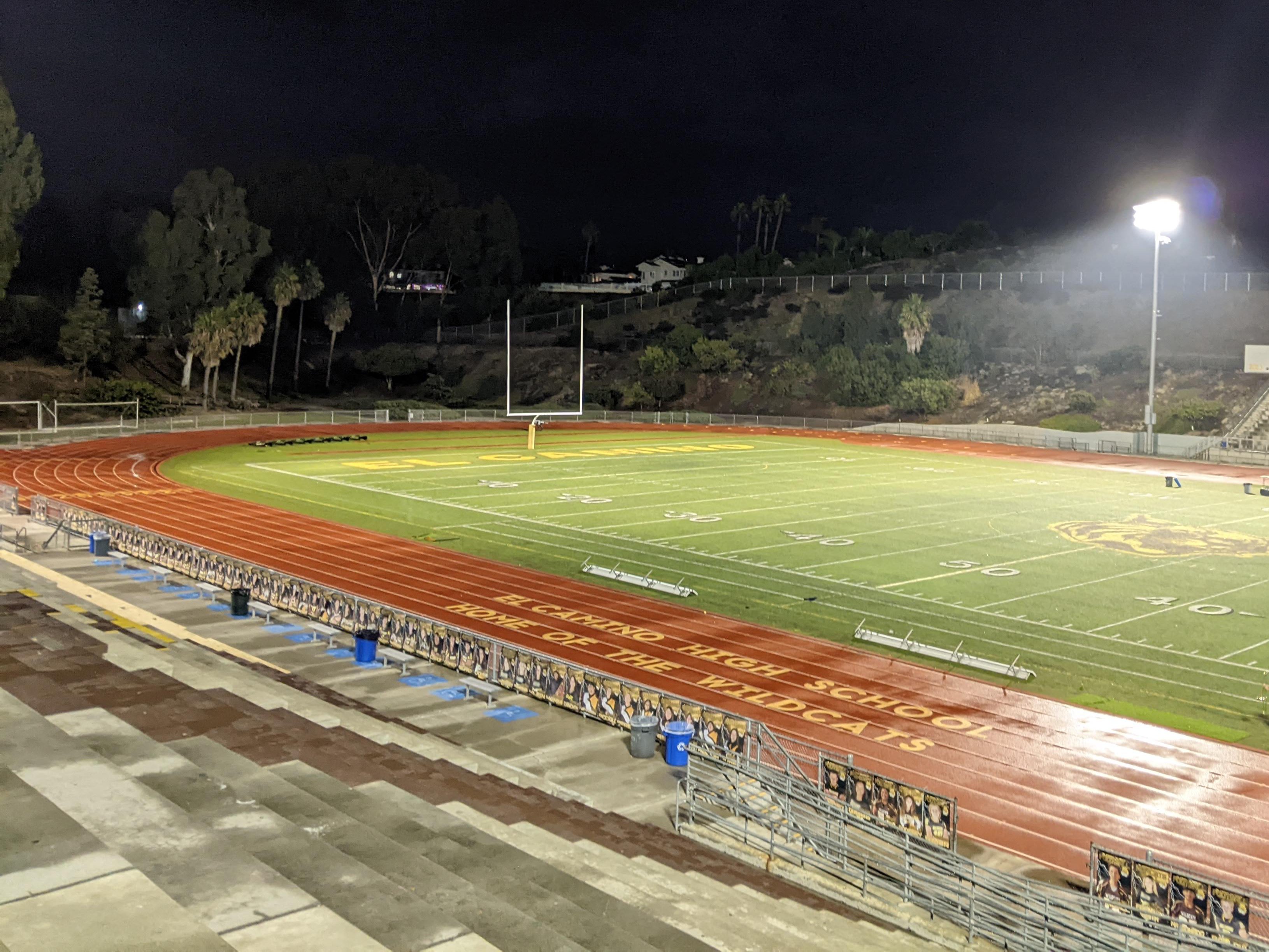 High school football stadium at night with lit artificial turf field and red running track.