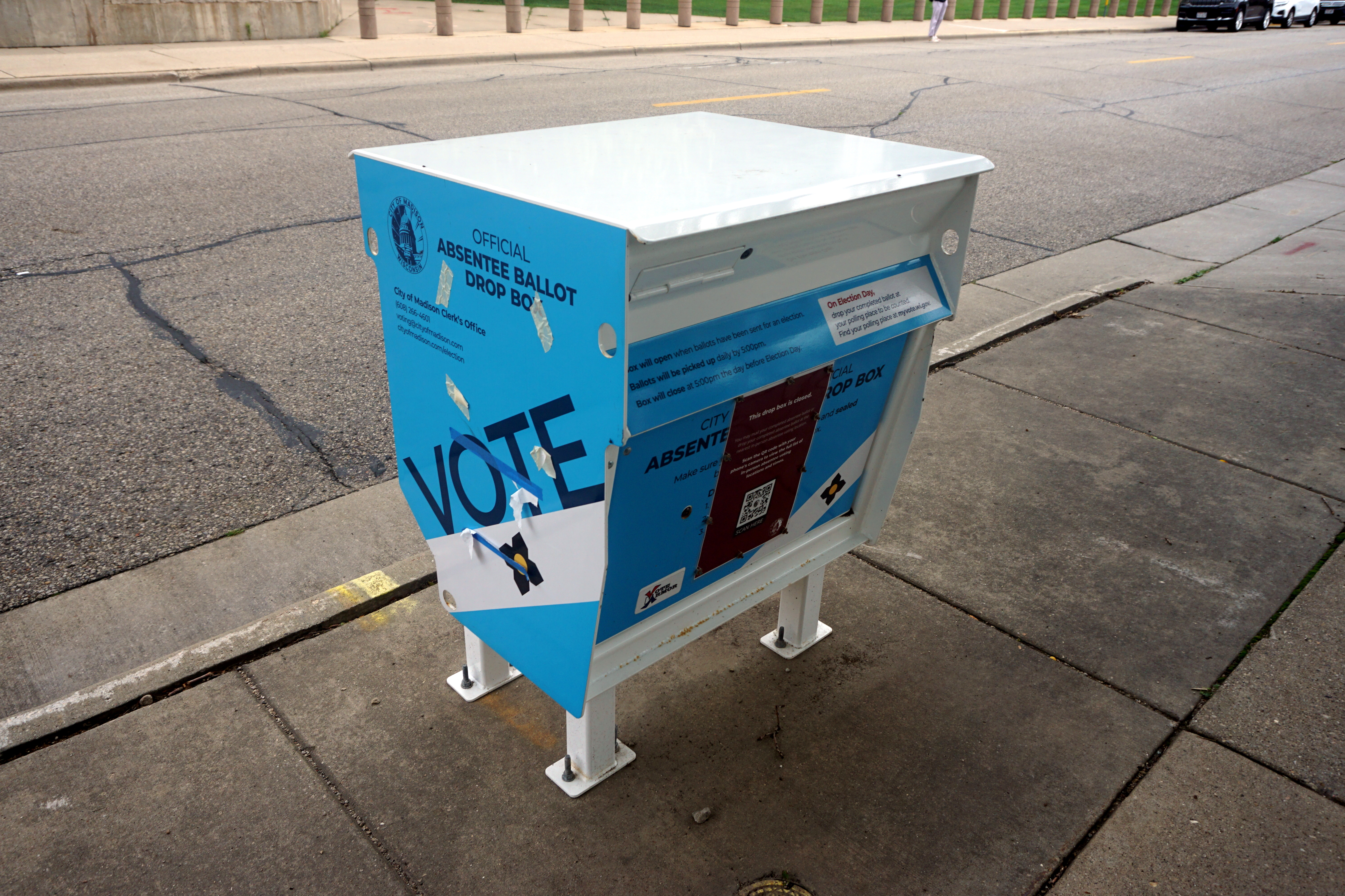 A blue and white official absentee ballot drop box on a concrete sidewalk.