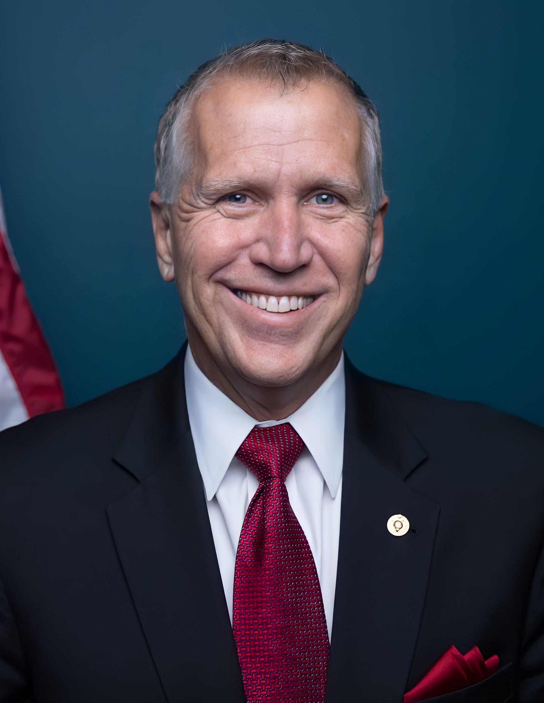 Man in dark suit and red tie smiling before a teal background with an American flag.