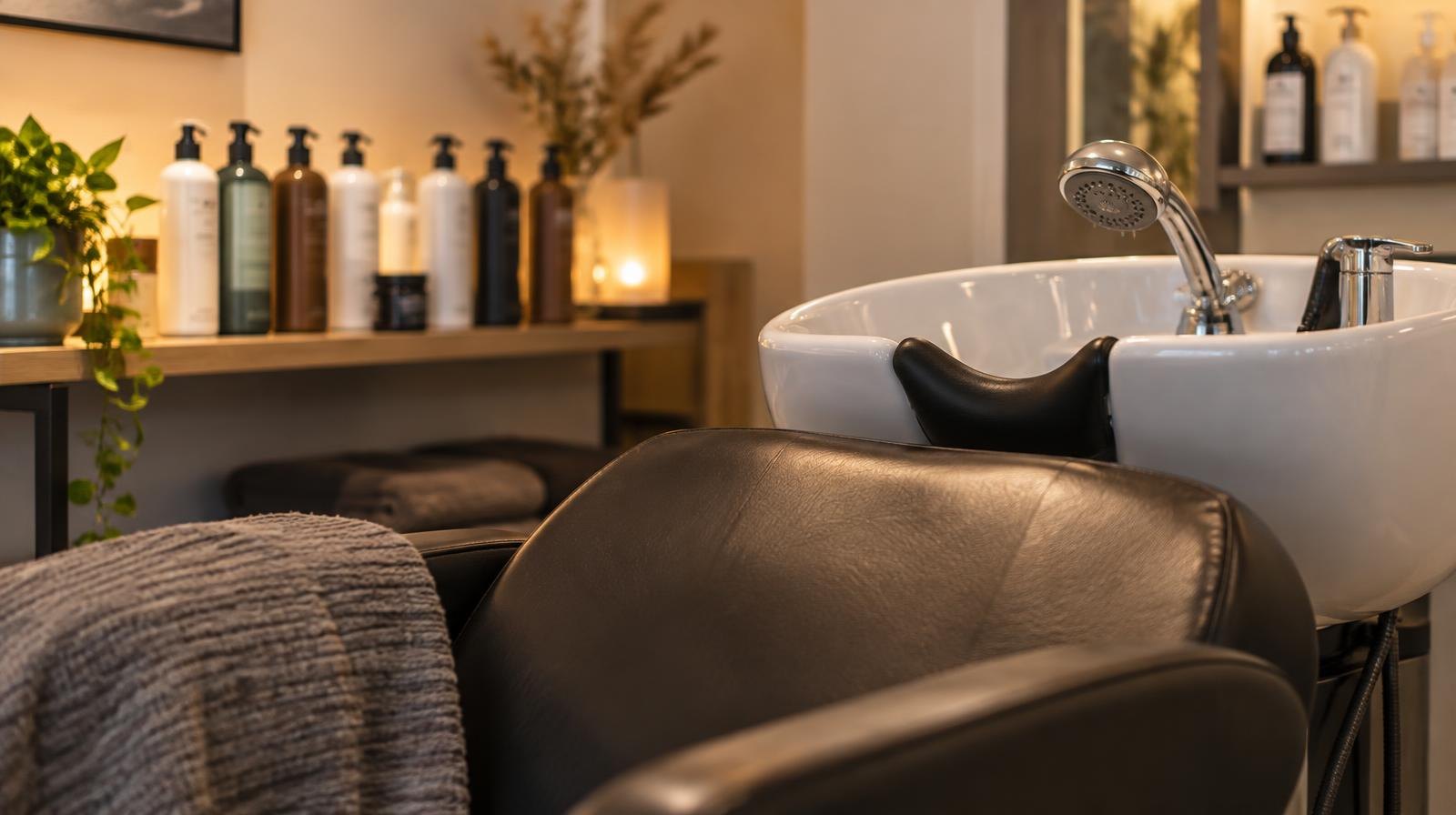 Reclined salon chair with showerhead attachment and hair care product bottles on a shelf.