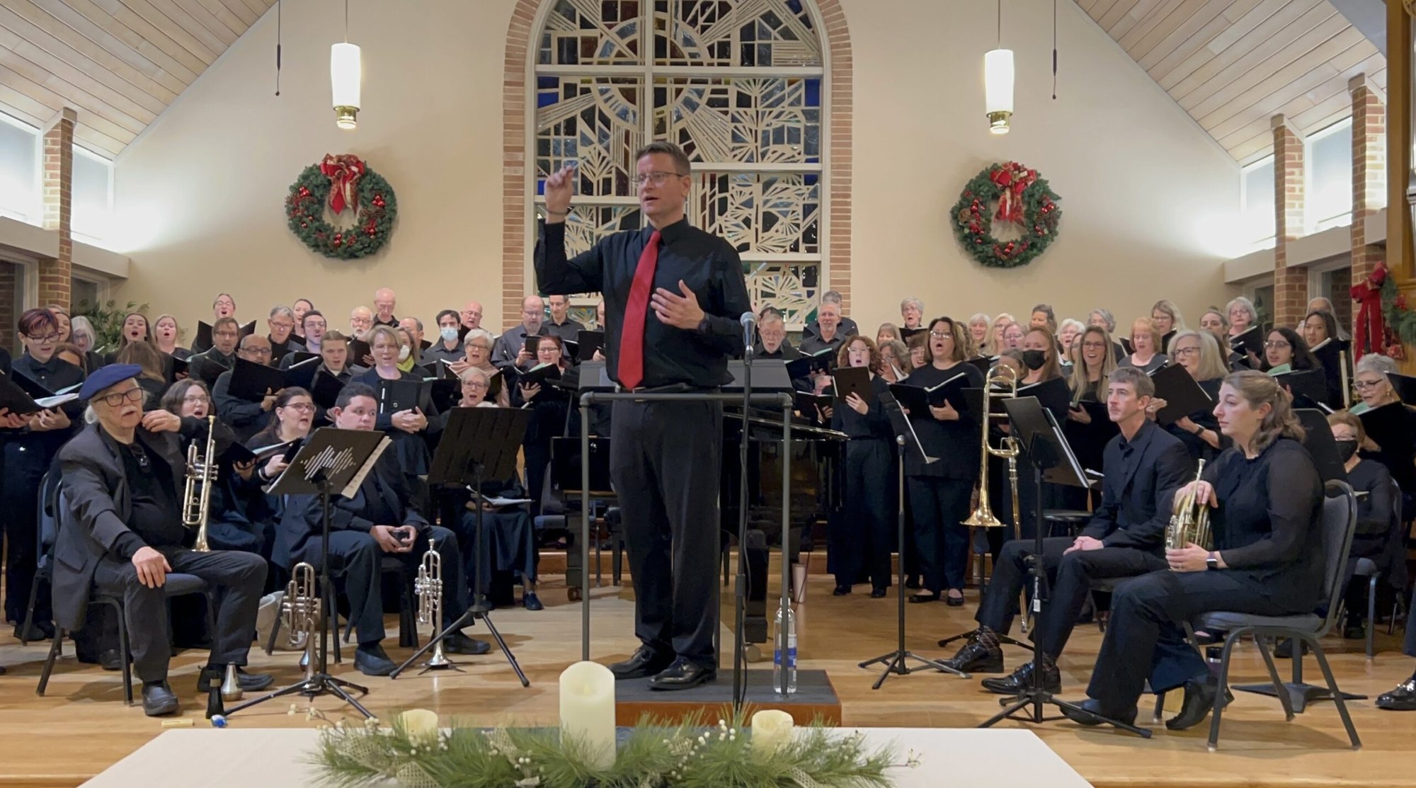 A conductor leads a large choir in black robes inside a church decorated with holiday wreaths.