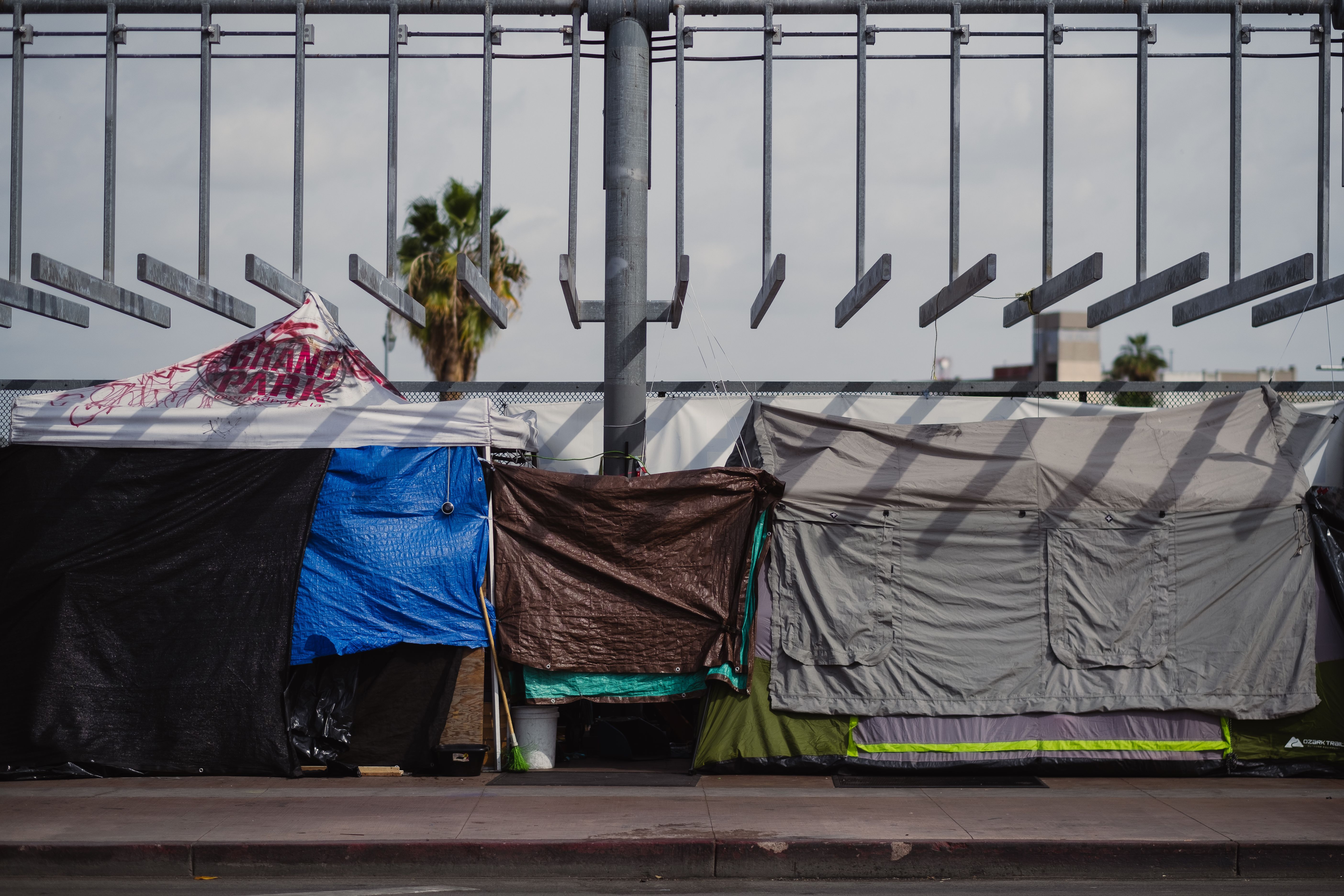Tents and tarps line a sidewalk beneath a freeway overpass in an urban area.