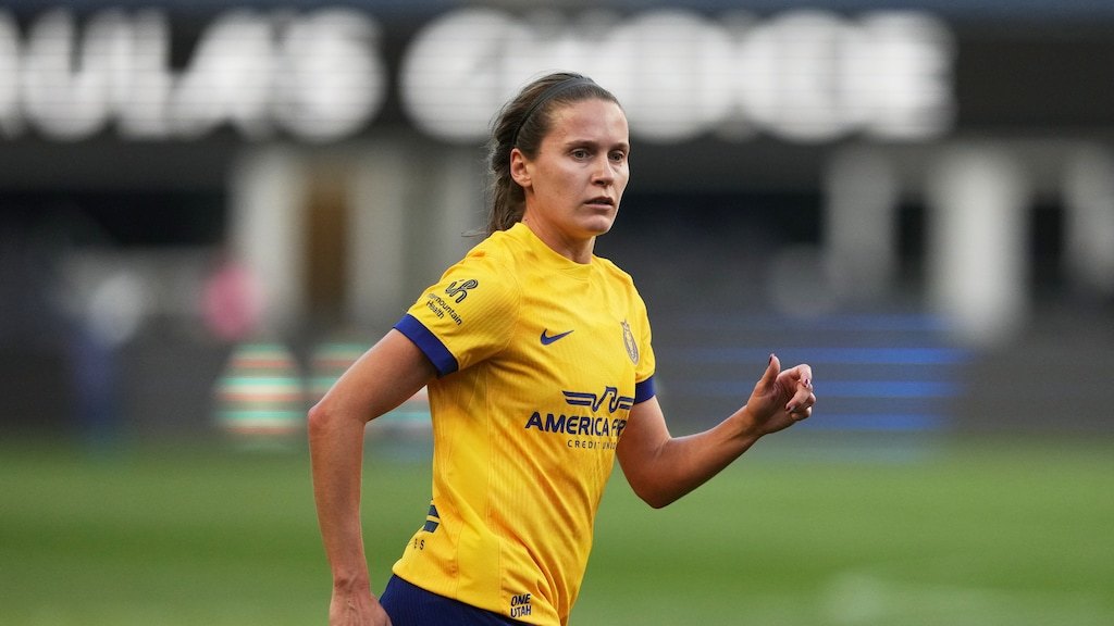 Female soccer player in yellow jersey jogging on a grass field with stadium in background.