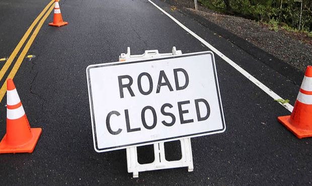 A white Road Closed sign on a barricade with two orange traffic cones on an asphalt road.