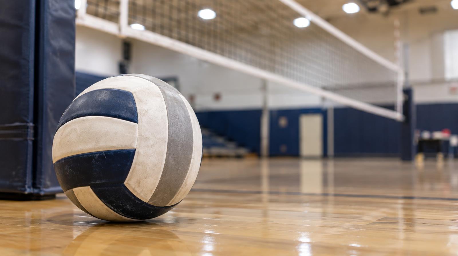 A volleyball resting on a hardwood gym floor near a net post.