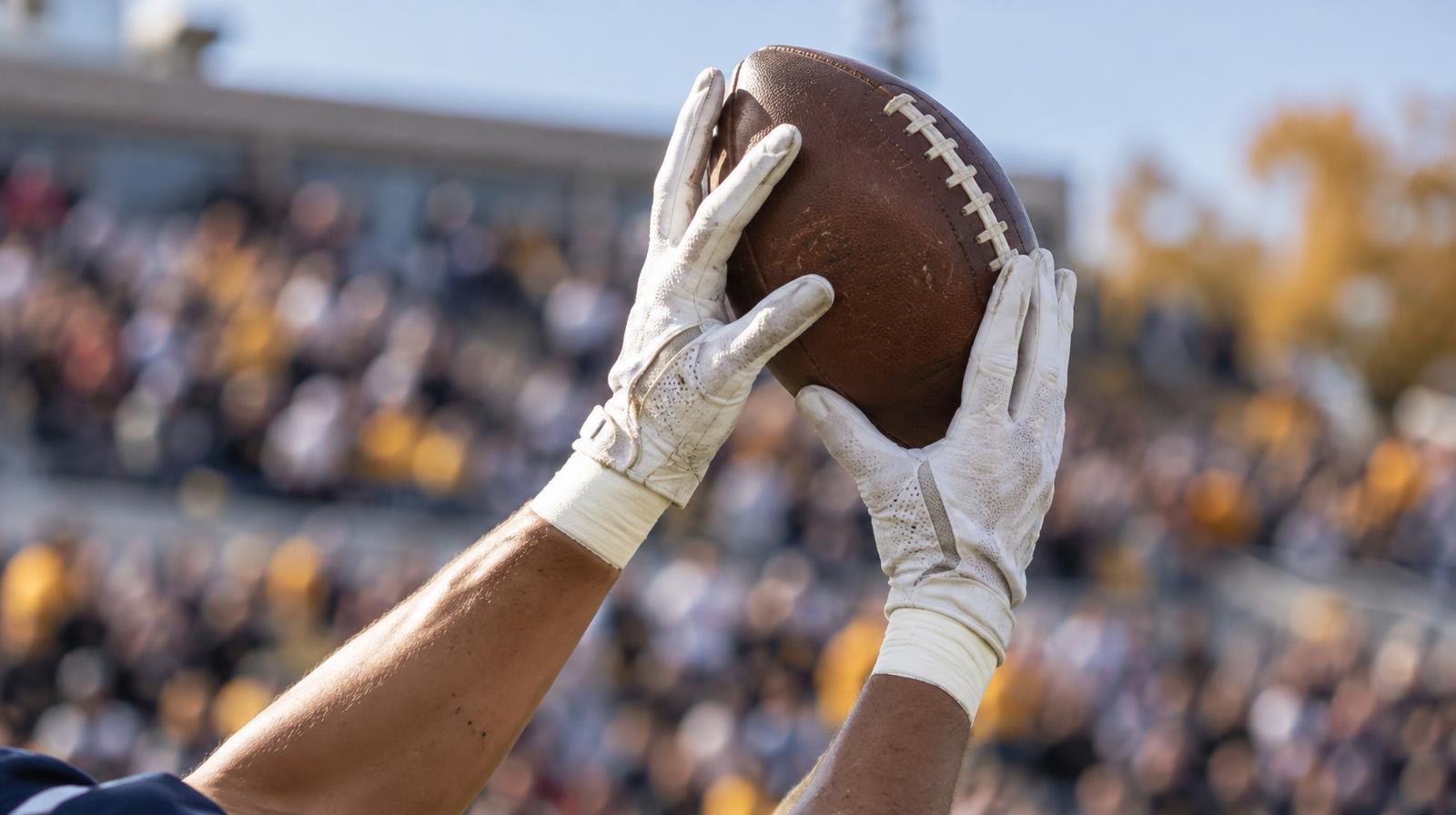Football receiver's gloves catching a spiral pass with a blurred stadium crowd behind.