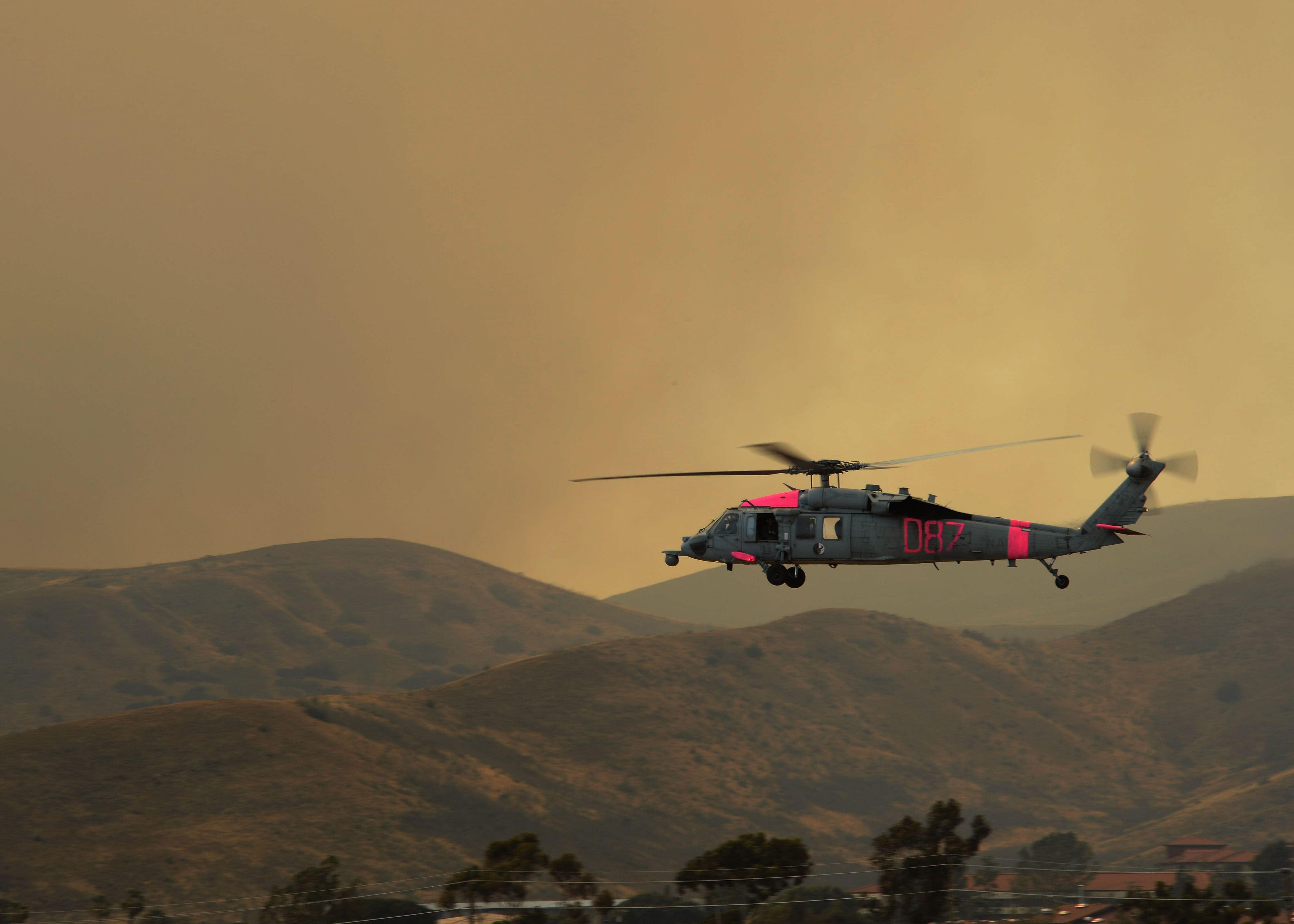 Military helicopter in flight against a hazy, smoke-filled orange sky over rolling hills.