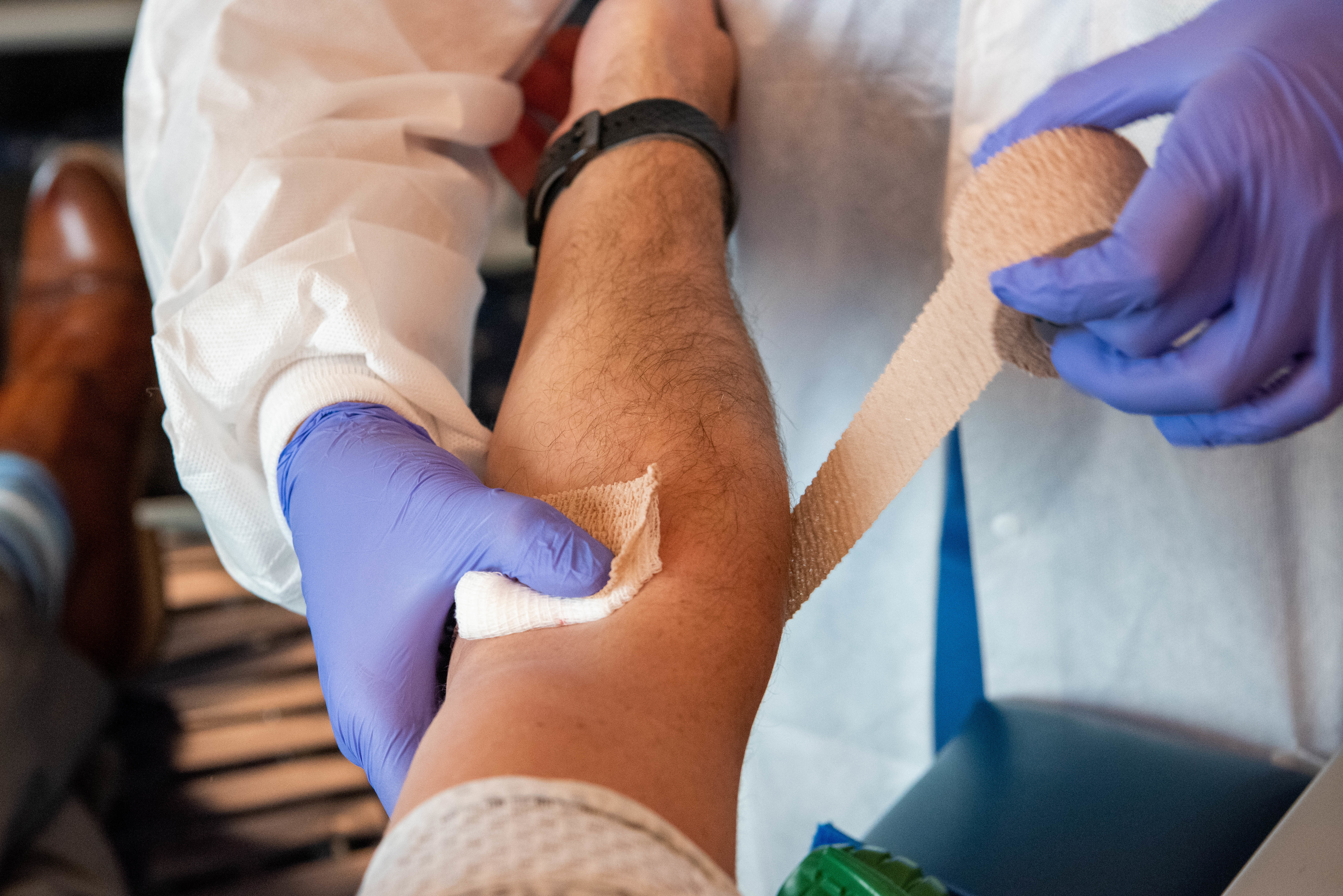 Gloved hands wrap a bandage around a donor's arm at the blood draw site.