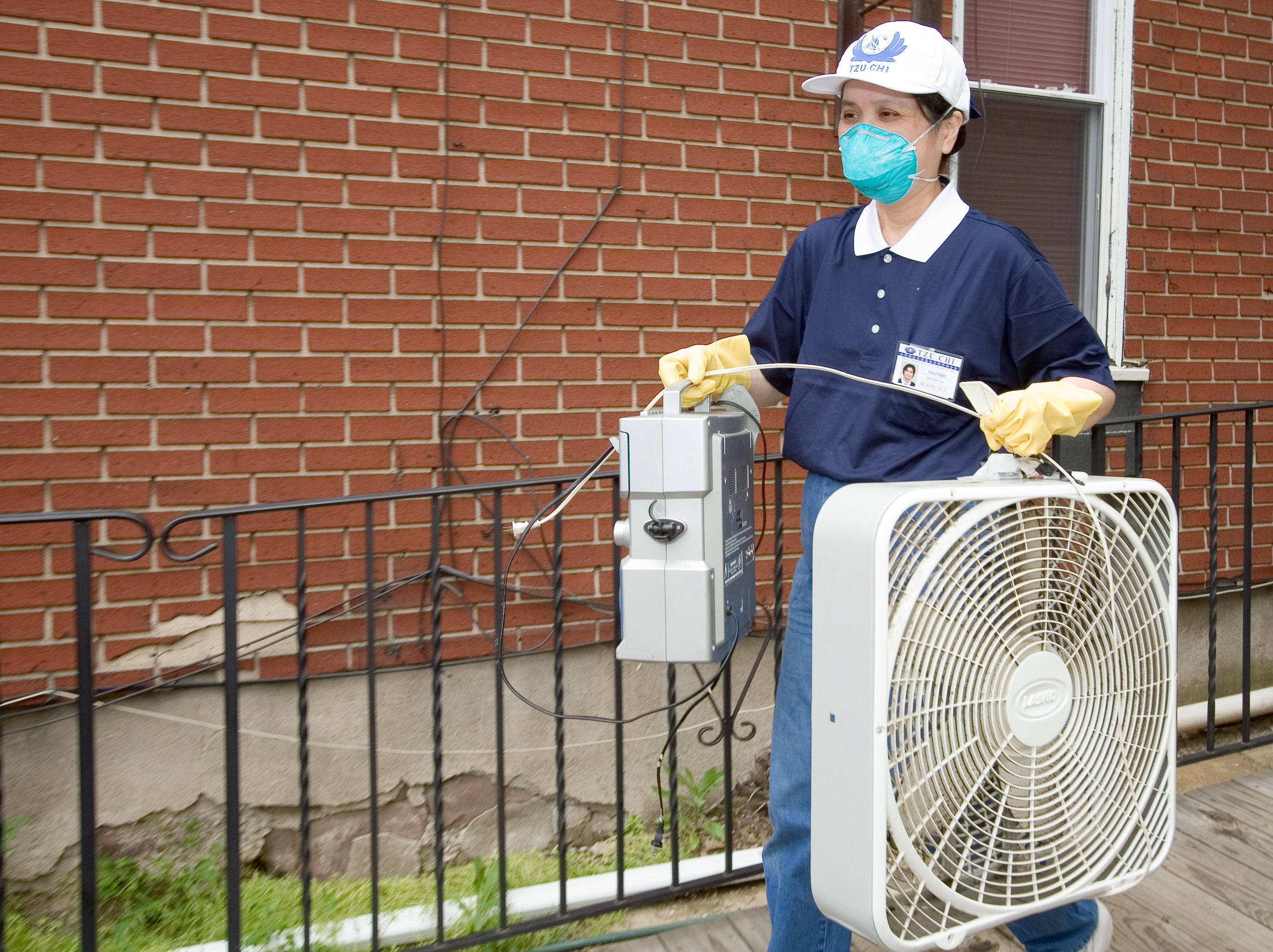 A person in a mask and yellow rubber gloves carries a box fan outside a brick building.
