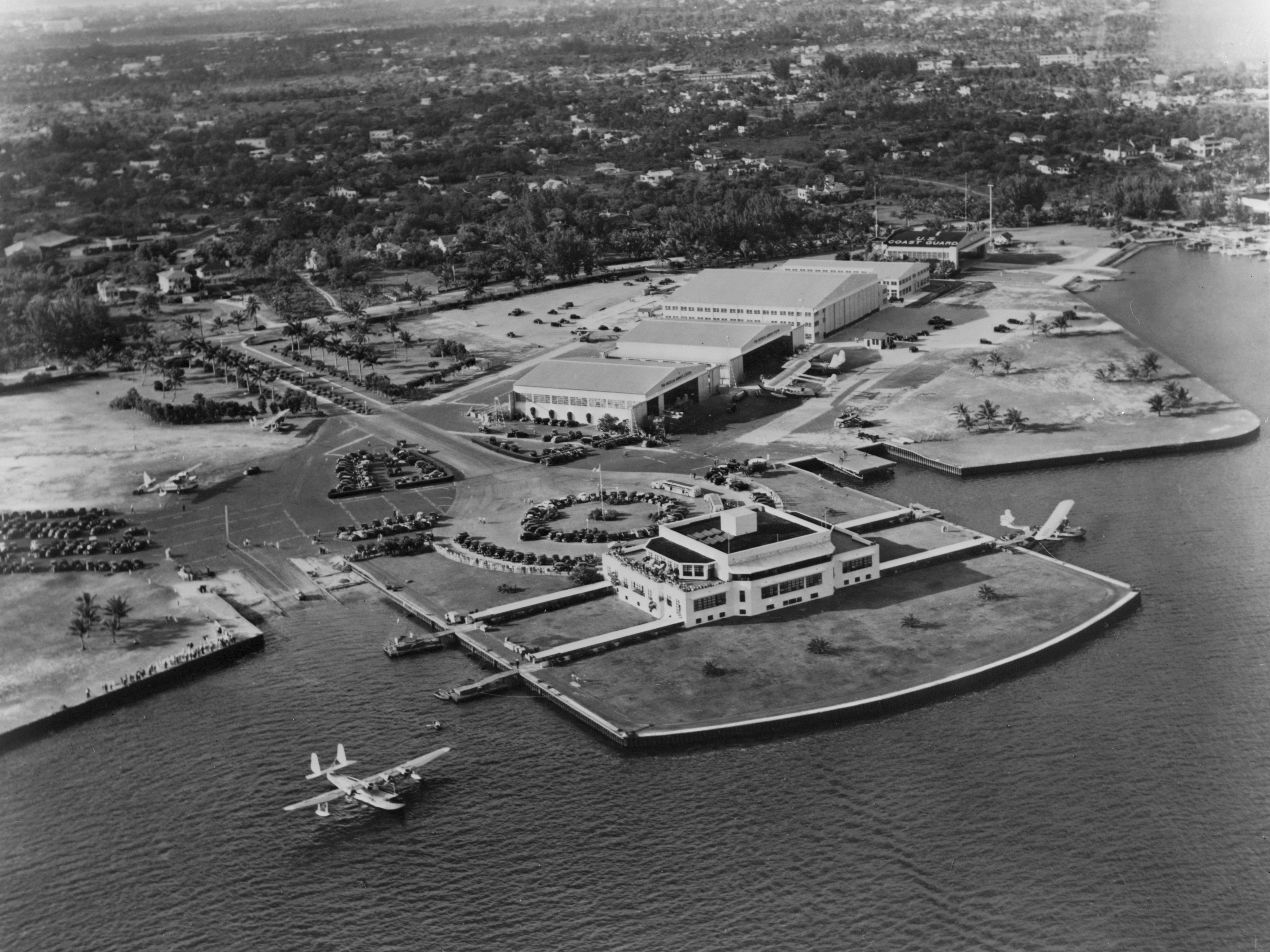 Aerial black-and-white photo of a waterfront terminal building with seaplanes on the water.