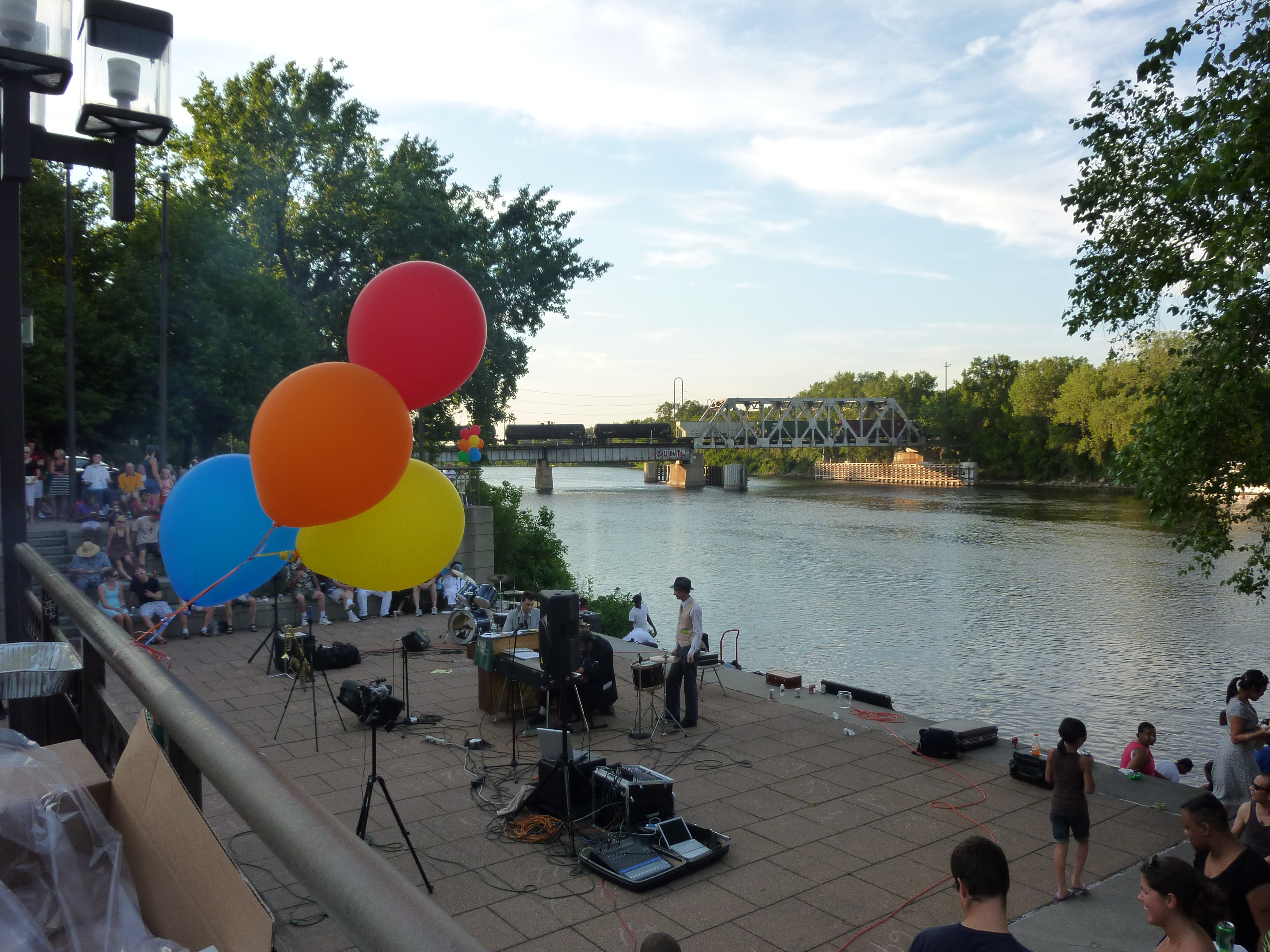 Crowd gathered at an outdoor community event with colorful balloons and a live music stage at dusk.