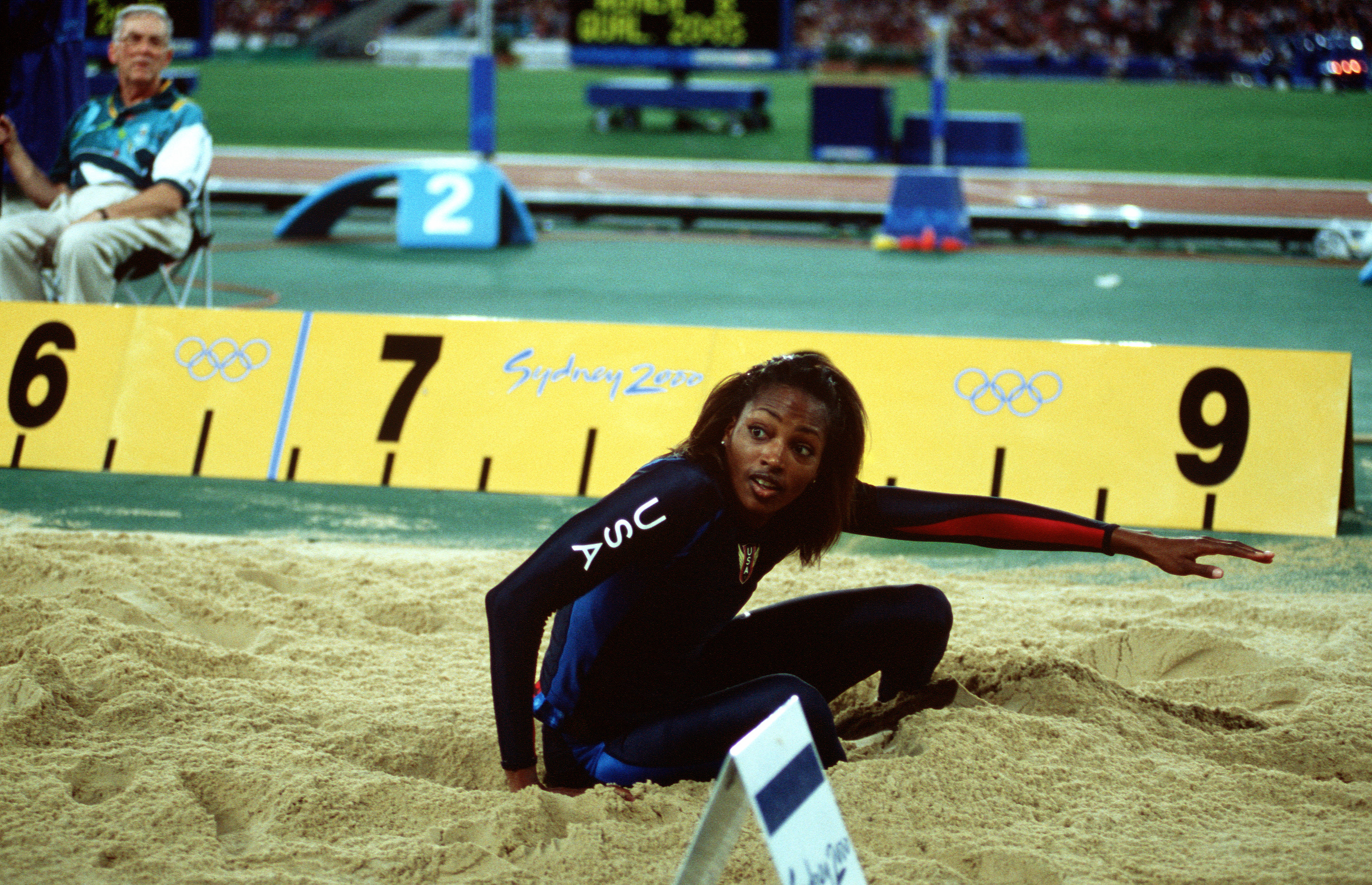 Female athlete in a USA uniform crouched in a long jump sandpit, arms extended.