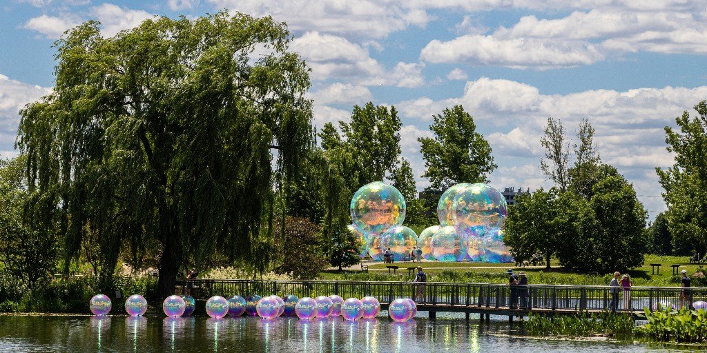 Large iridescent bubble sculptures installed along a lake shore at a botanical garden with visitors walking nearby.