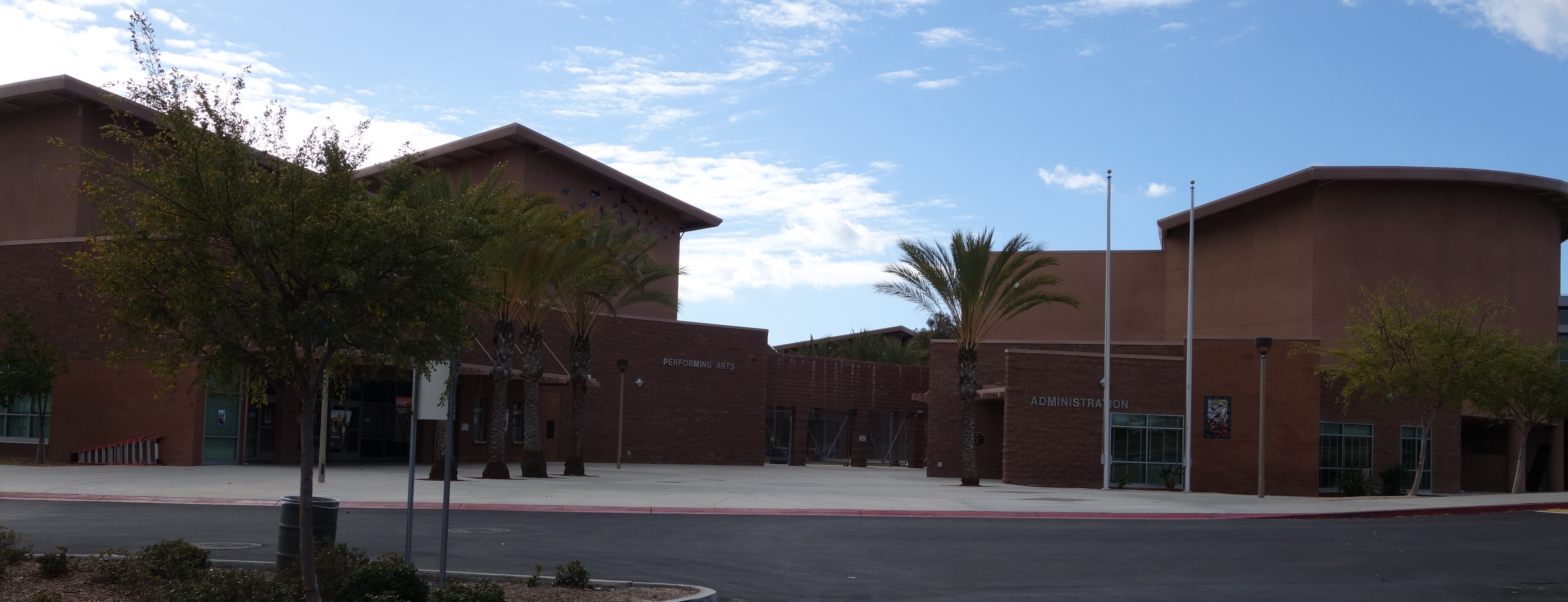 Exterior front entrance of a Southwest-style school building with palm trees and flagpoles.