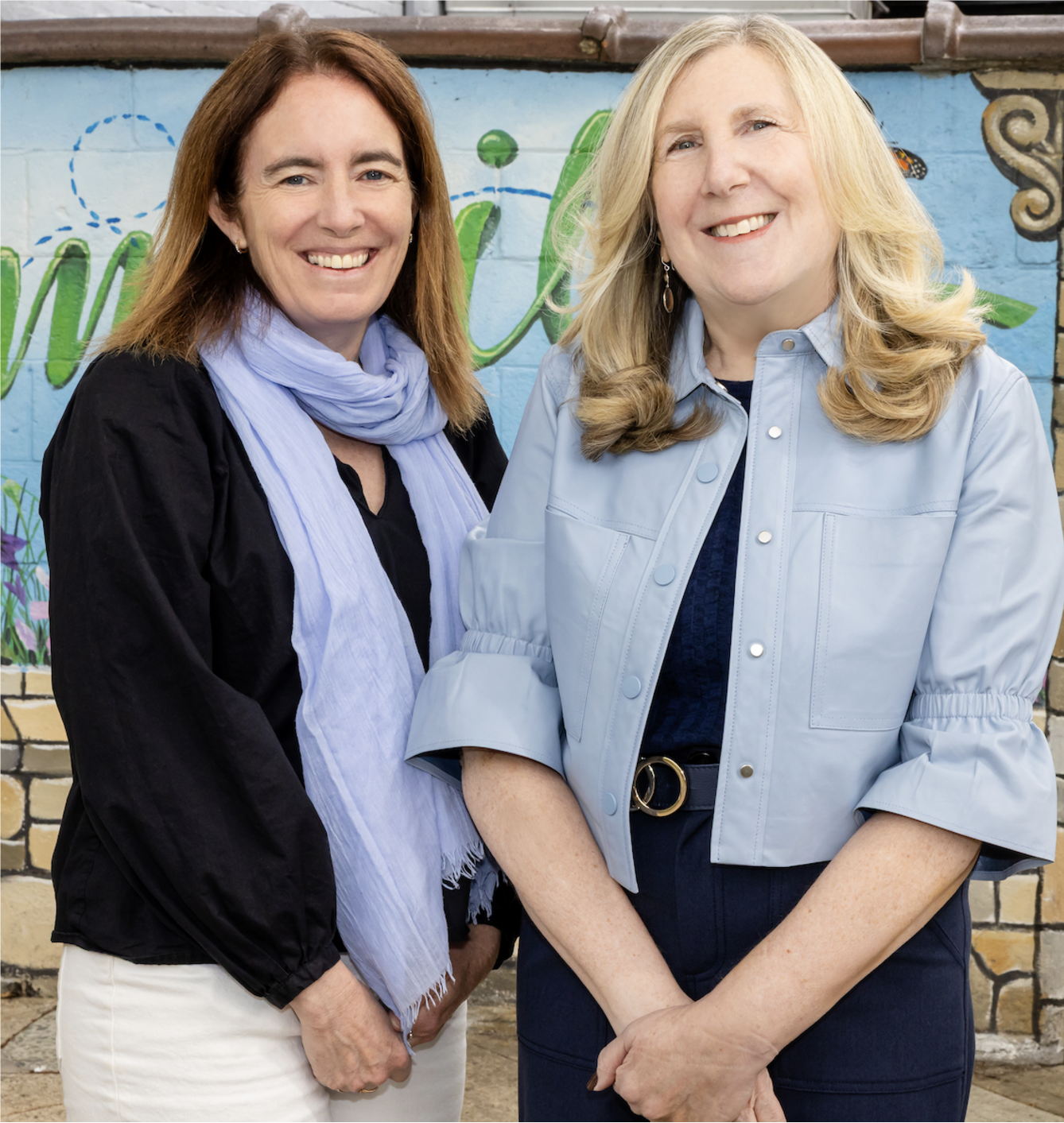 Two smiling women stand side by side in front of a colorful painted mural.