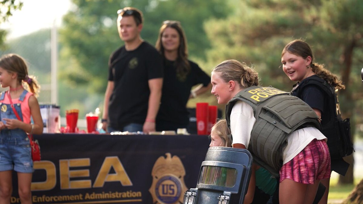 A police officer in a tactical vest crouches near children at an outdoor community event table.
