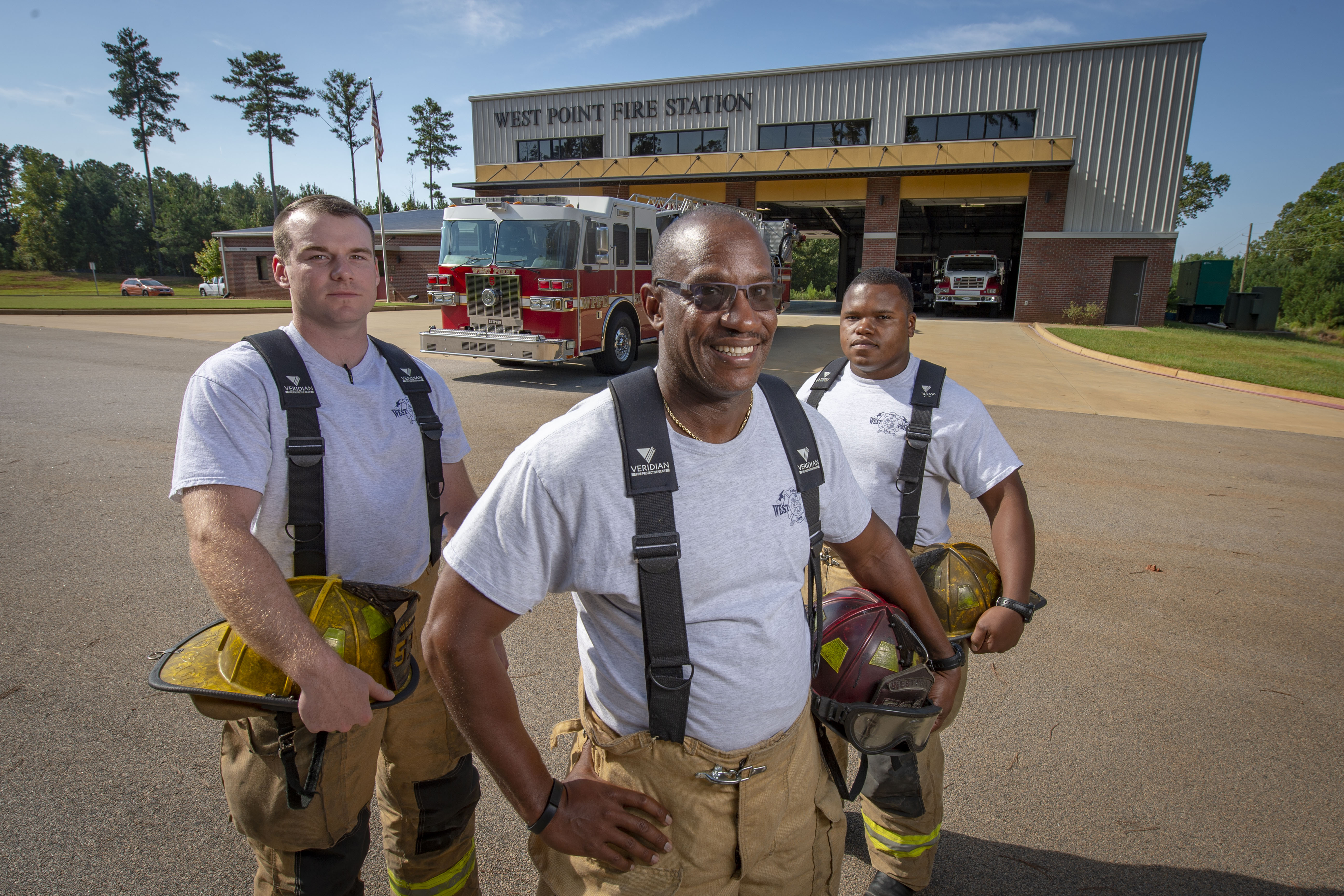 Three firefighters in turnout gear stand in front of a fire station with a red truck visible inside.