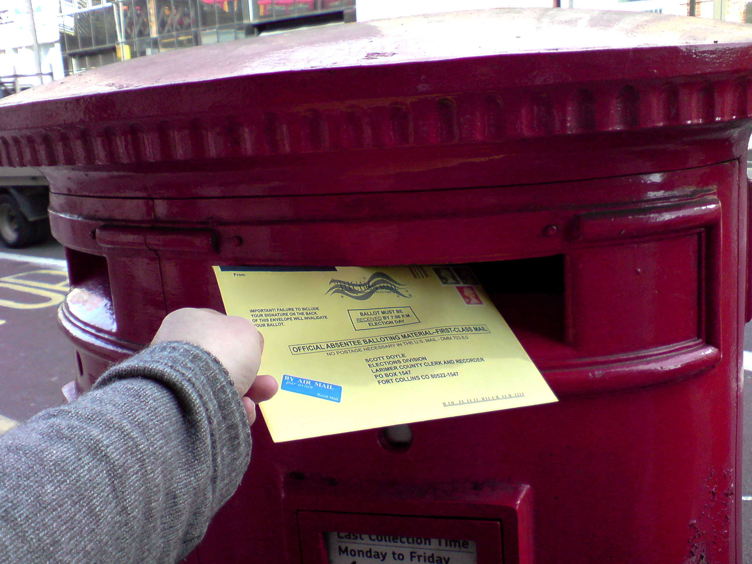 Hand inserting a yellow official absentee ballot envelope into a red post box.