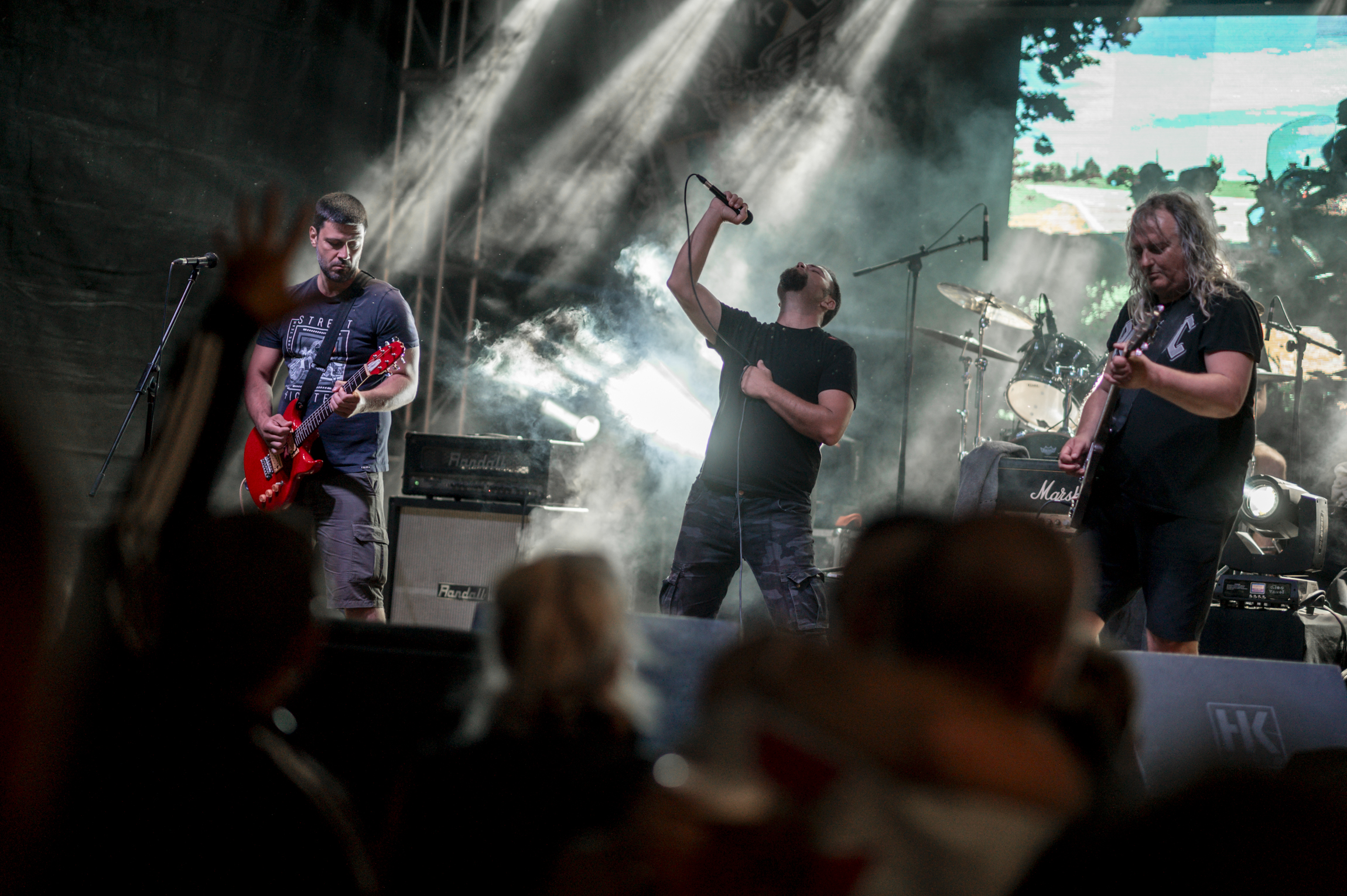 Three musicians perform on a brightly lit outdoor stage before a crowd.