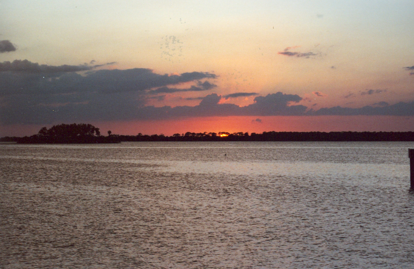 Sunset over calm water with wooden dock pilings and a distant shoreline.