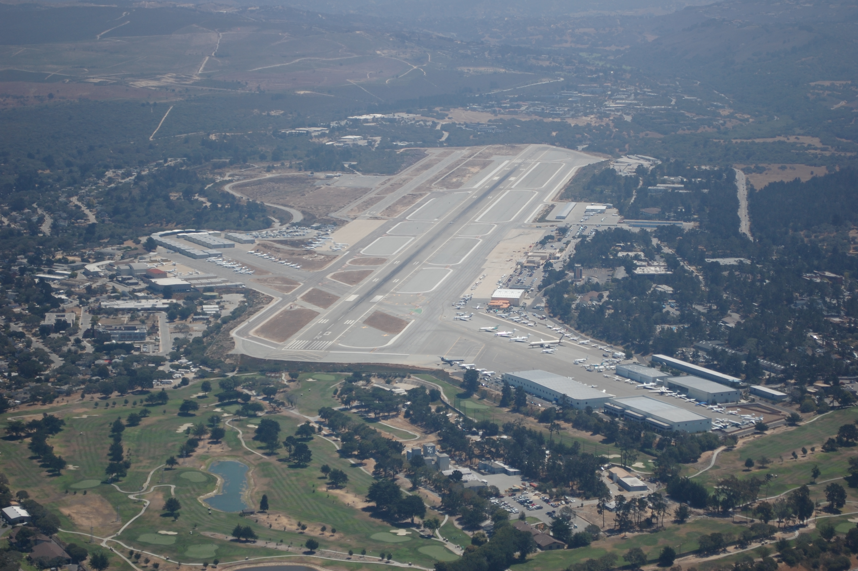 Aerial view of an airport with a single runway, terminal buildings, and parked aircraft.