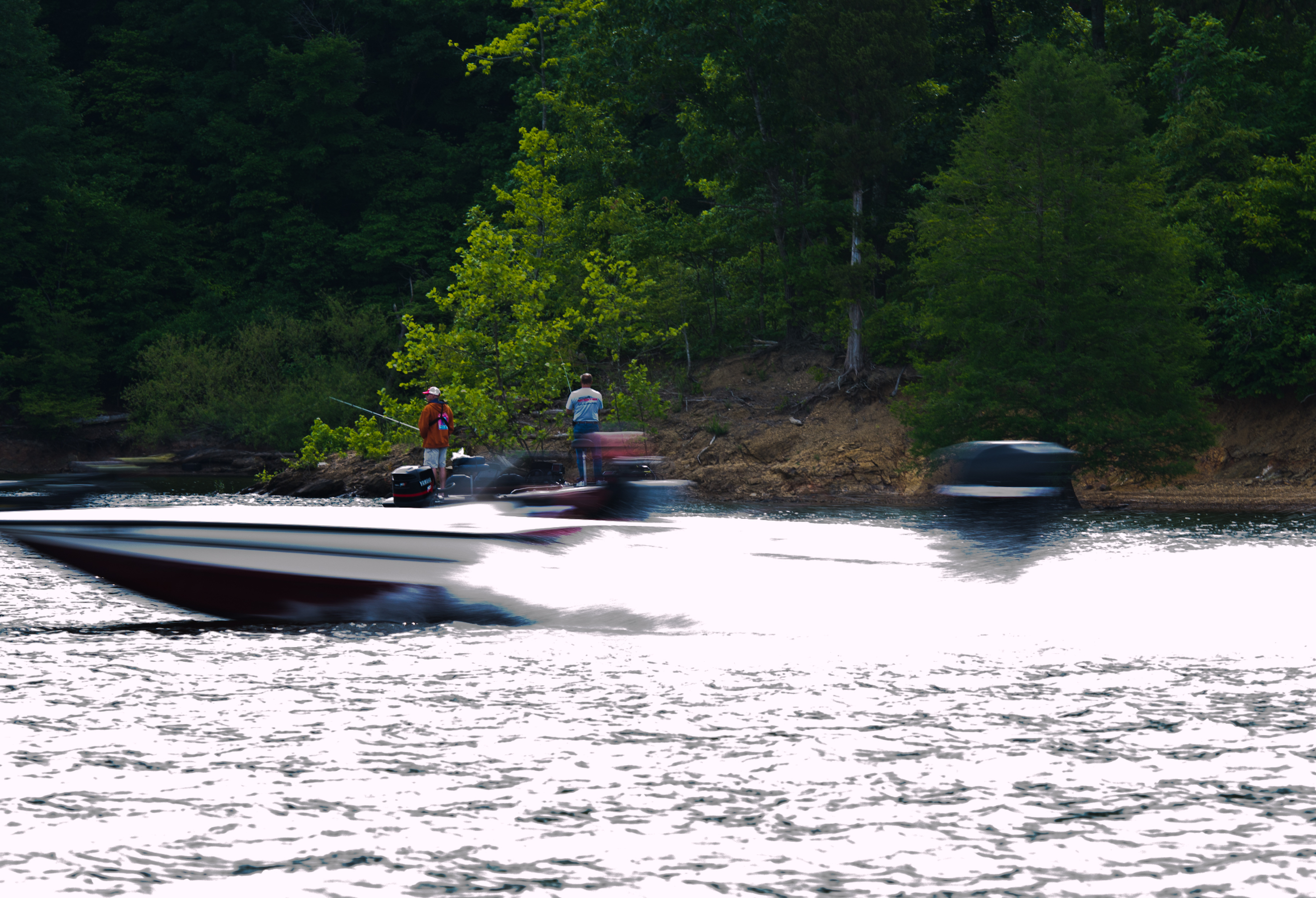 A bass boat speeds across a lake with two anglers standing on a nearby boat.