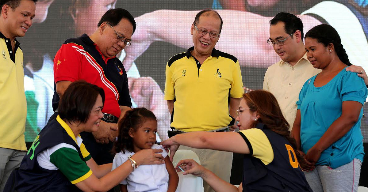 A young girl receives a vaccination injection from a health worker while adults look on.
