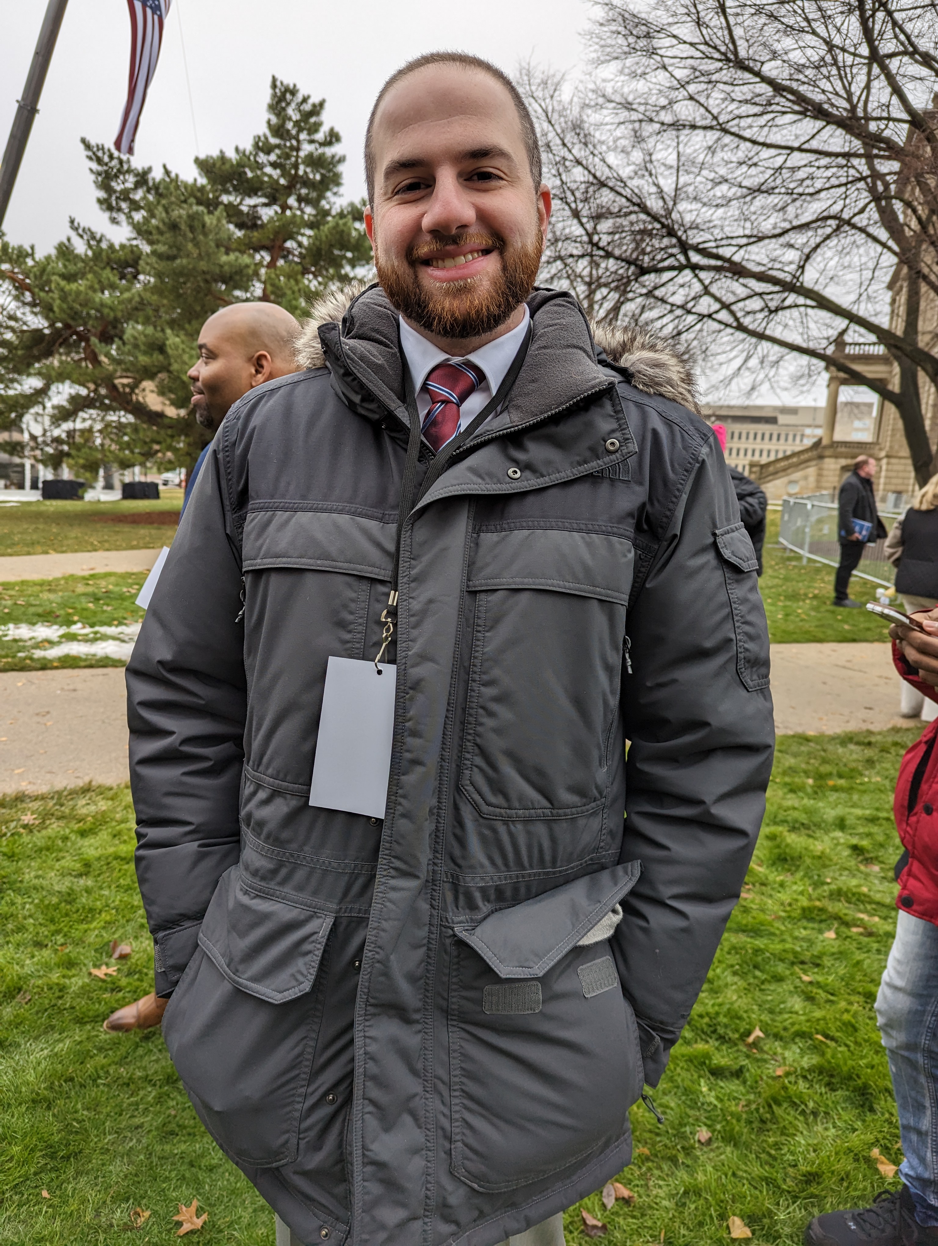Smiling man in a gray winter coat with a credential lanyard at an outdoor public event.