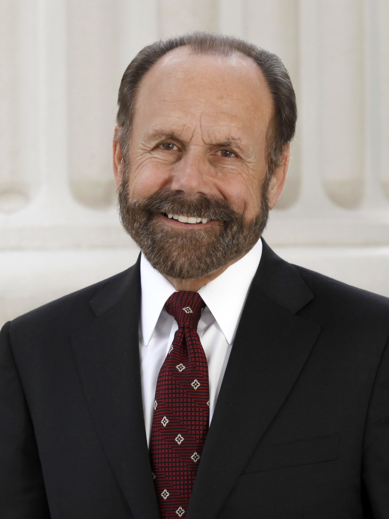 A smiling man with a gray beard wearing a dark suit and red patterned tie in front of white columns.