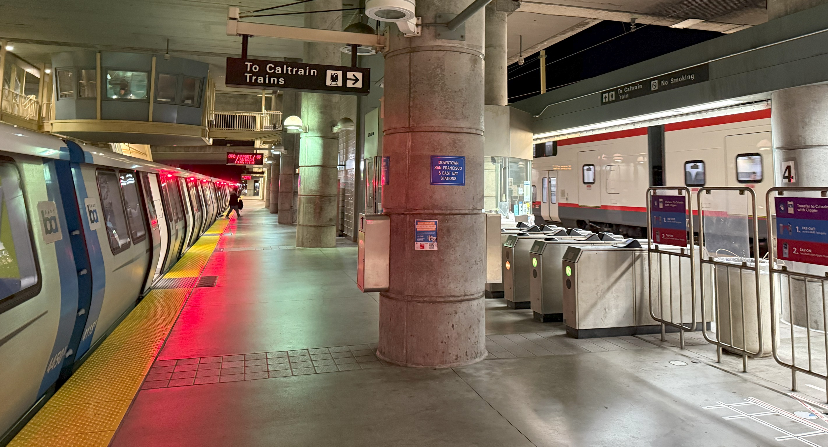 A BART train and a Caltrain train on opposite sides of a station platform with fare gates visible.