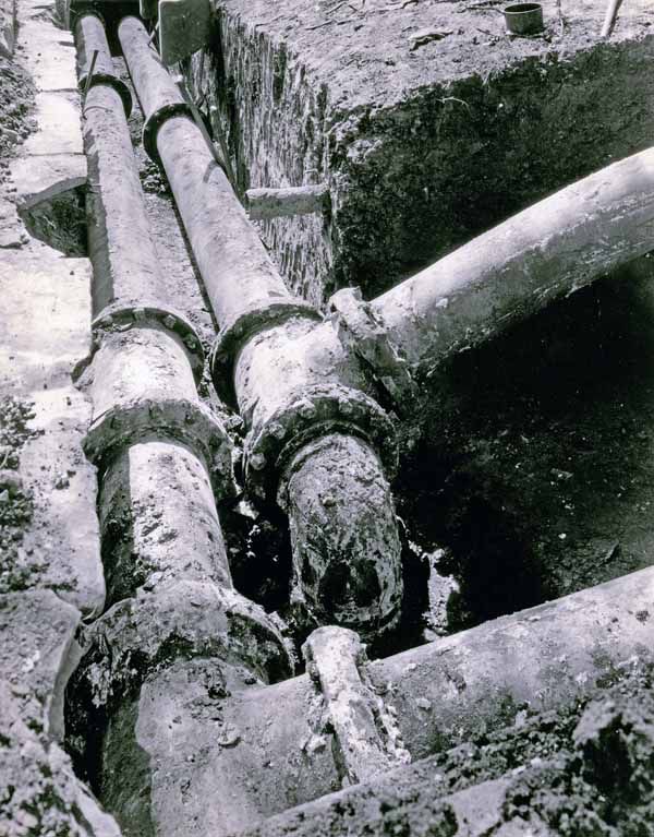 Two large corroded metal pipes exposed in an excavated earthen trench.