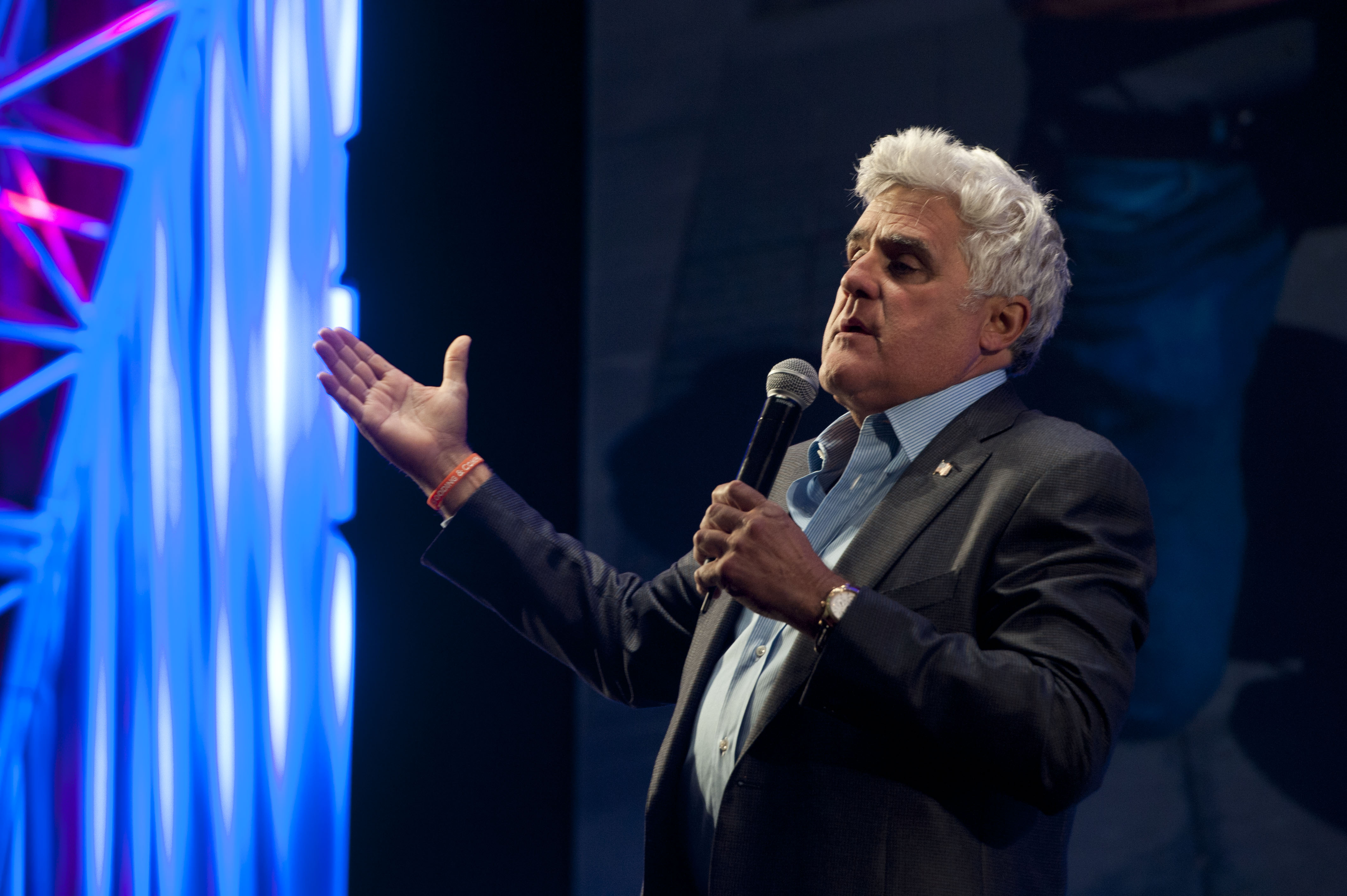 White-haired man speaks into a microphone on a stage, gesturing with one hand.