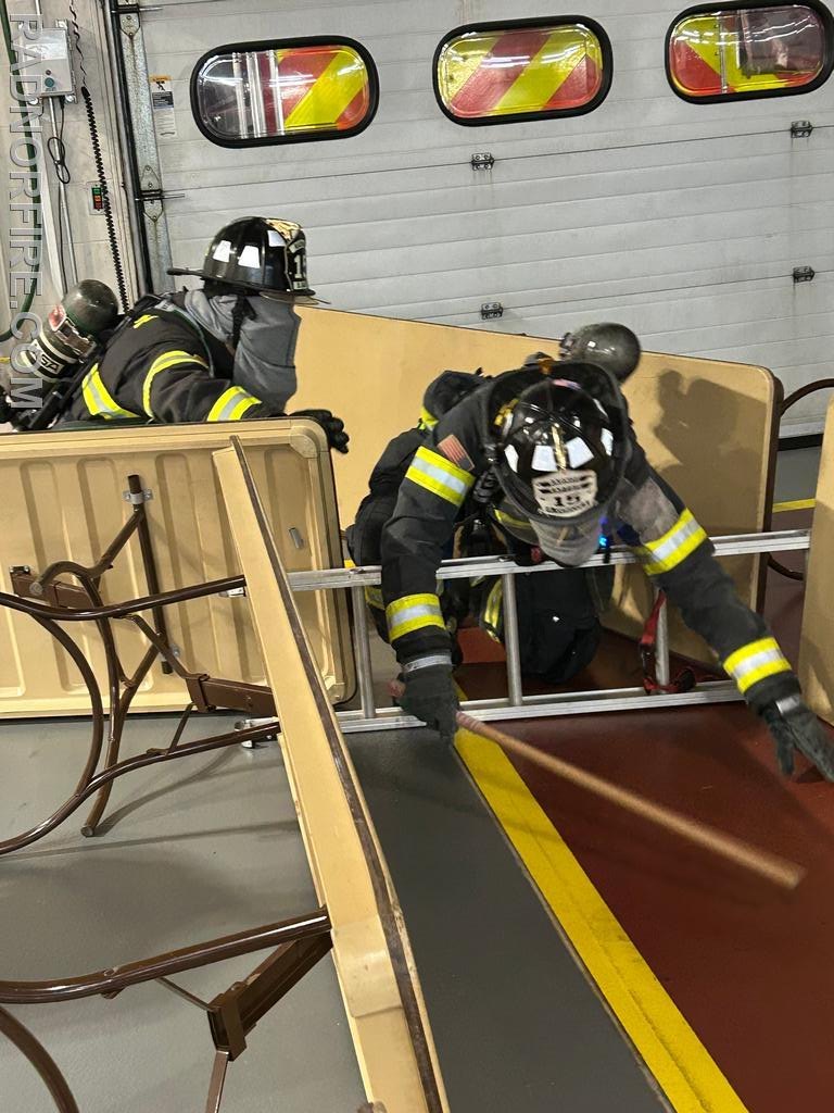 Two firefighters in full gear and blacked-out masks crawl through a narrow wooden obstacle course inside a fire station.