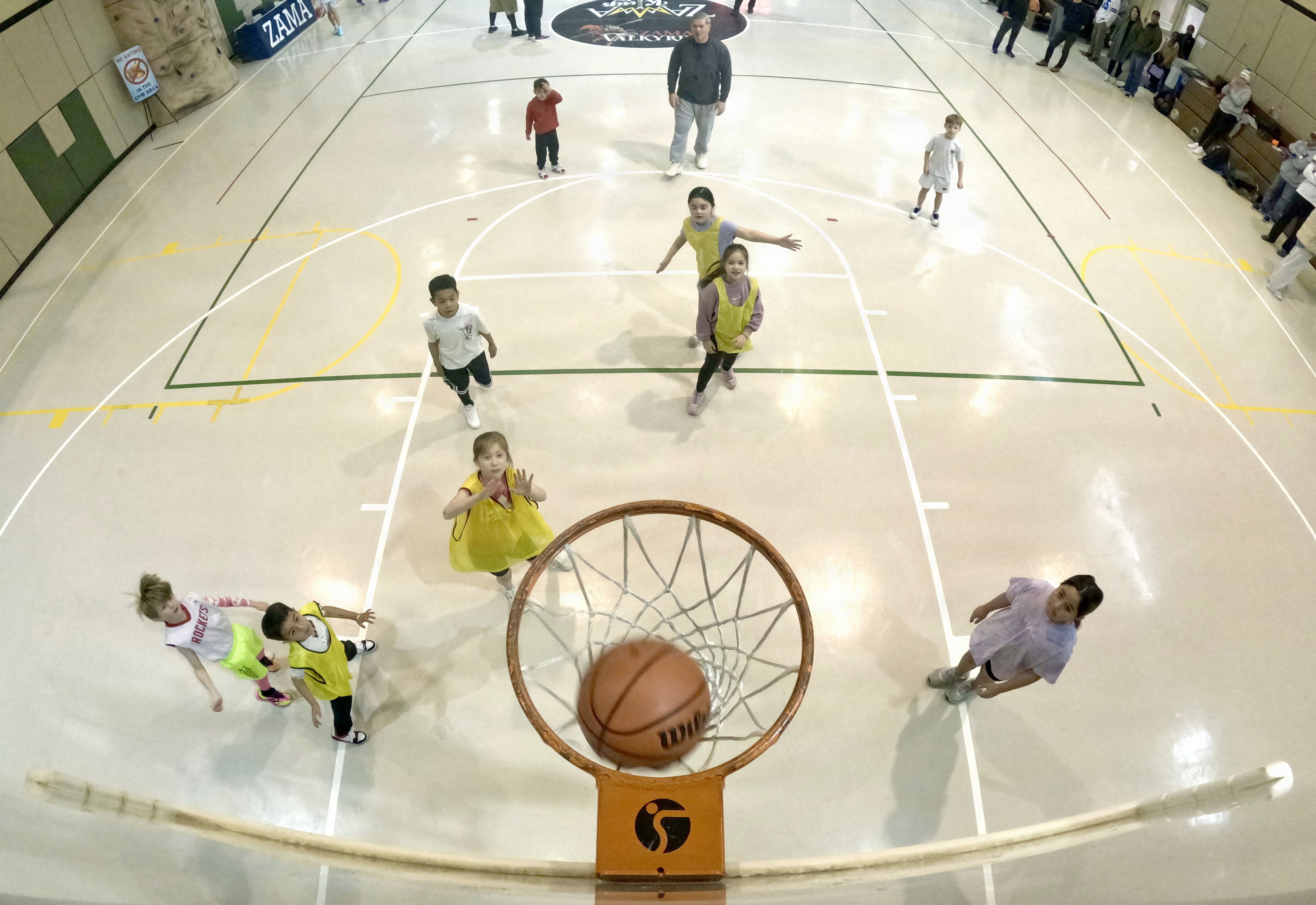 Children play a basketball scrimmage on an indoor court, viewed from above the hoop.