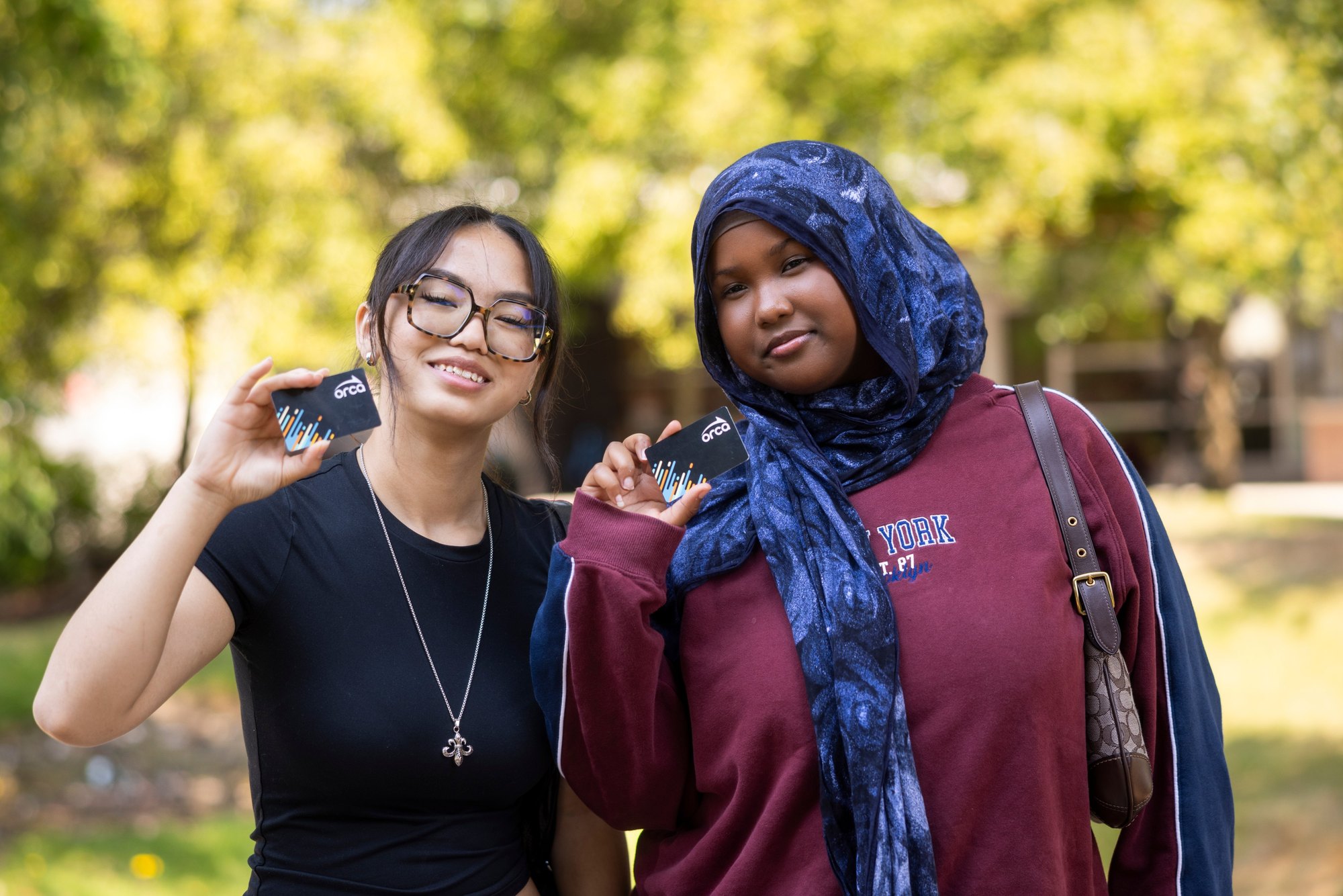 Two smiling young women outdoors each holding up a small dark transit pass card.