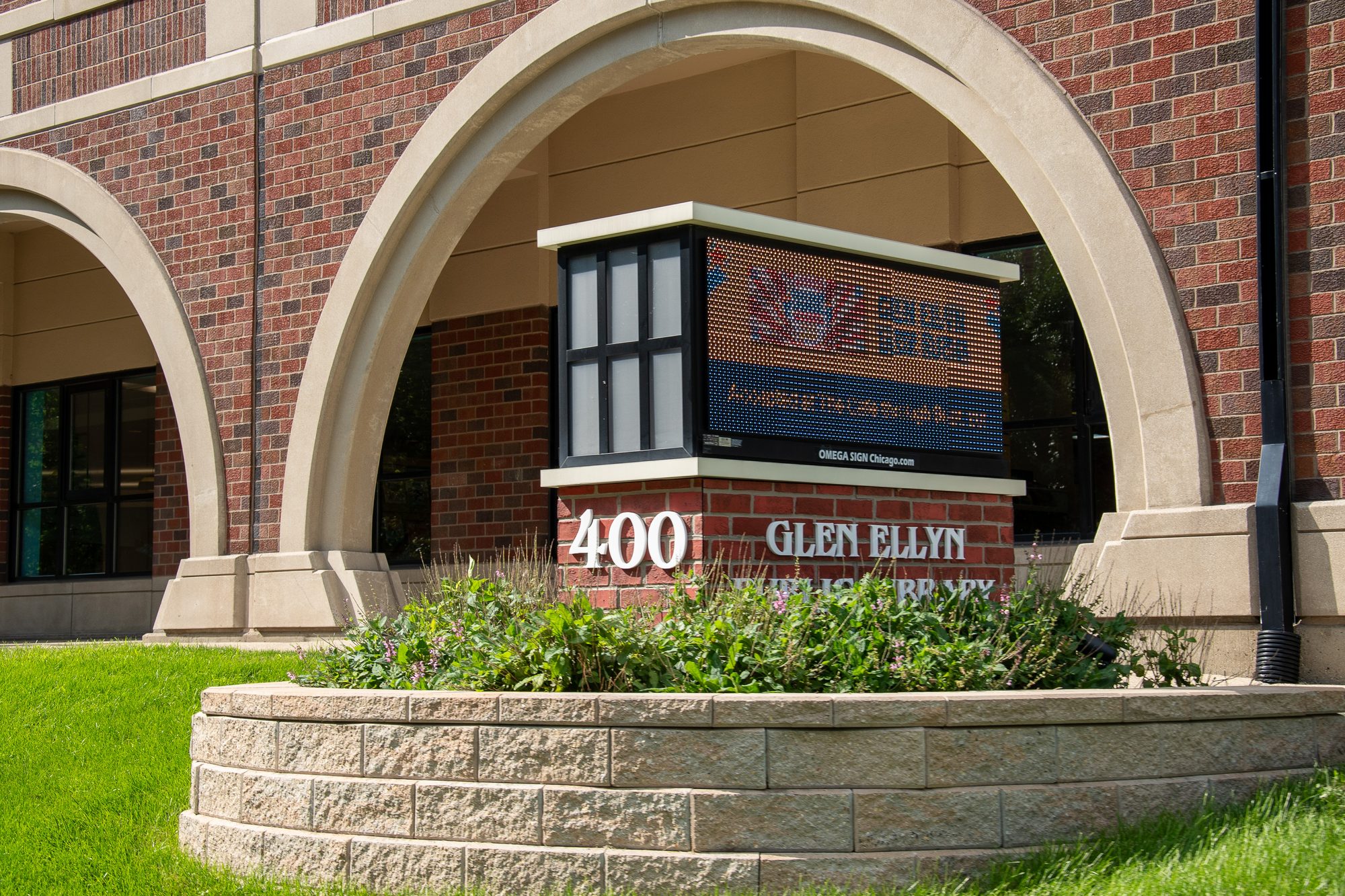 Brick public library exterior with arched entrance and landscaped sign out front.