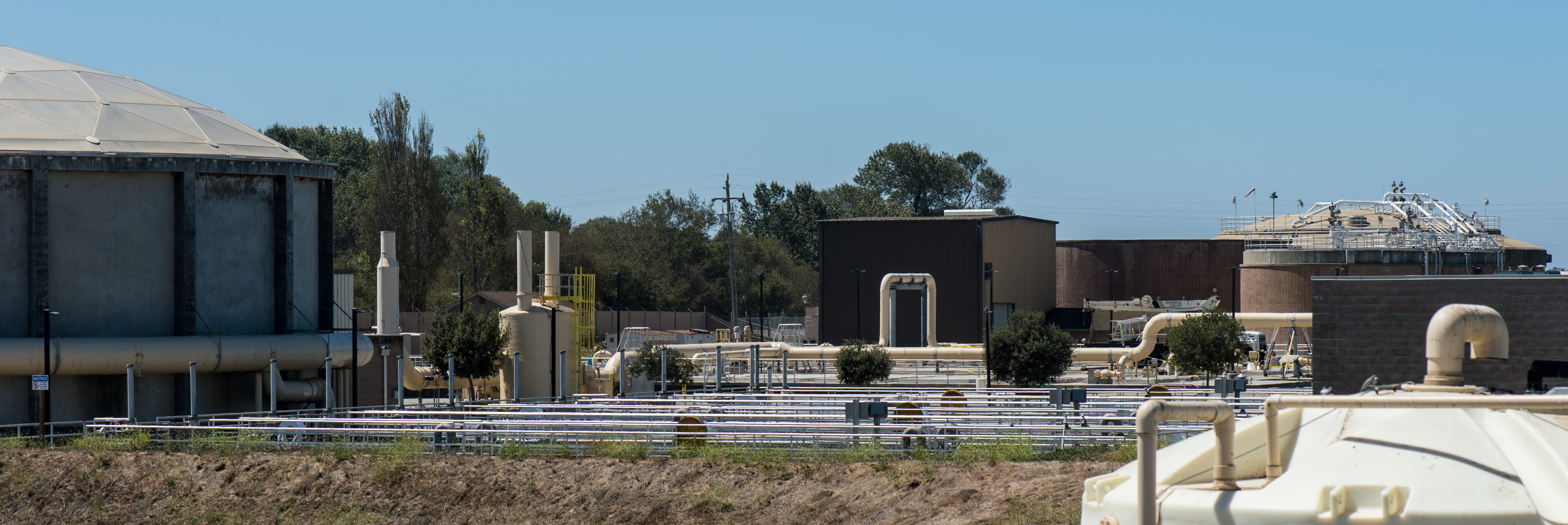 Industrial water treatment facility with pipes, tanks, and buildings under a clear blue sky.