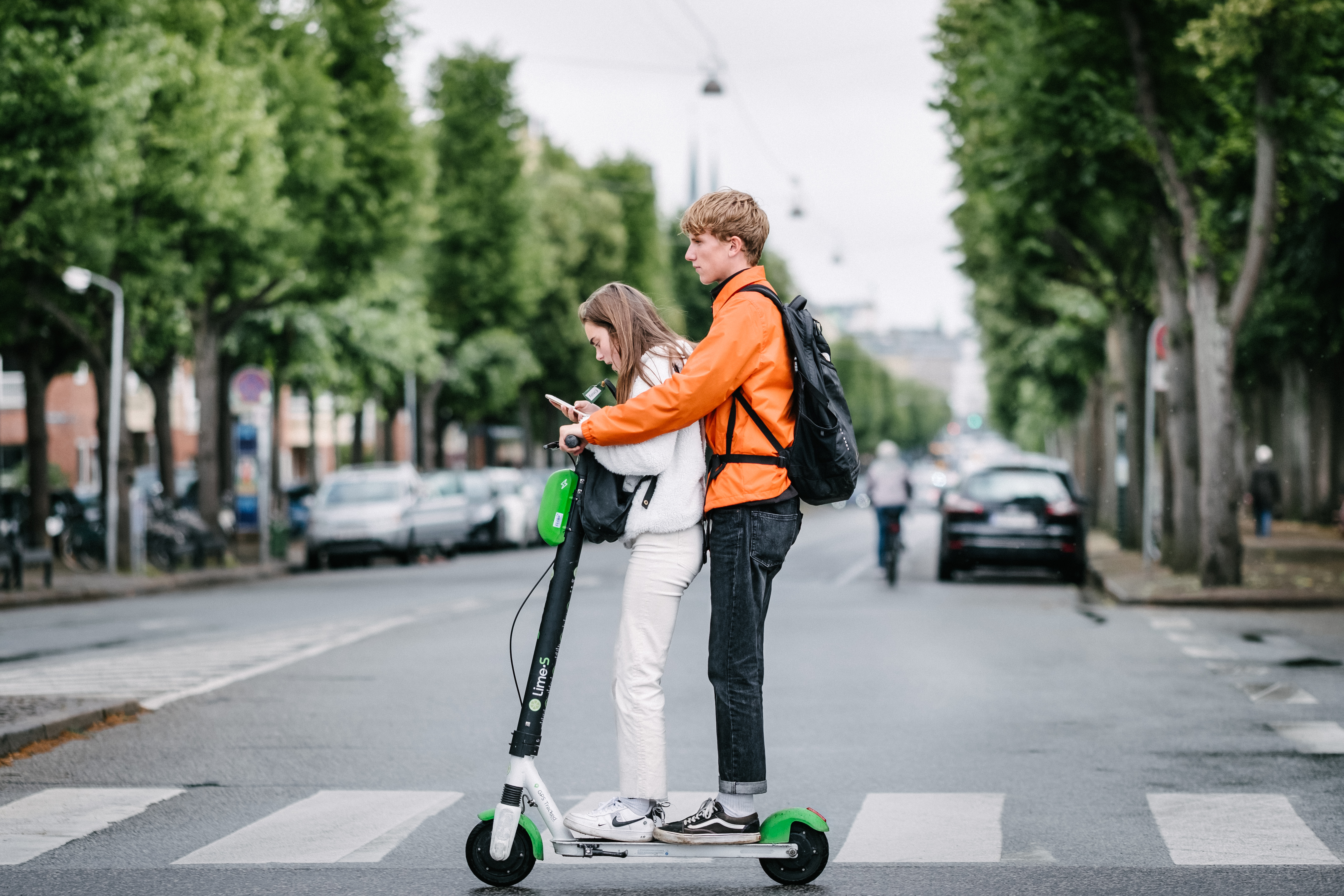 Two young people stopped on a green electric scooter at an urban crosswalk.