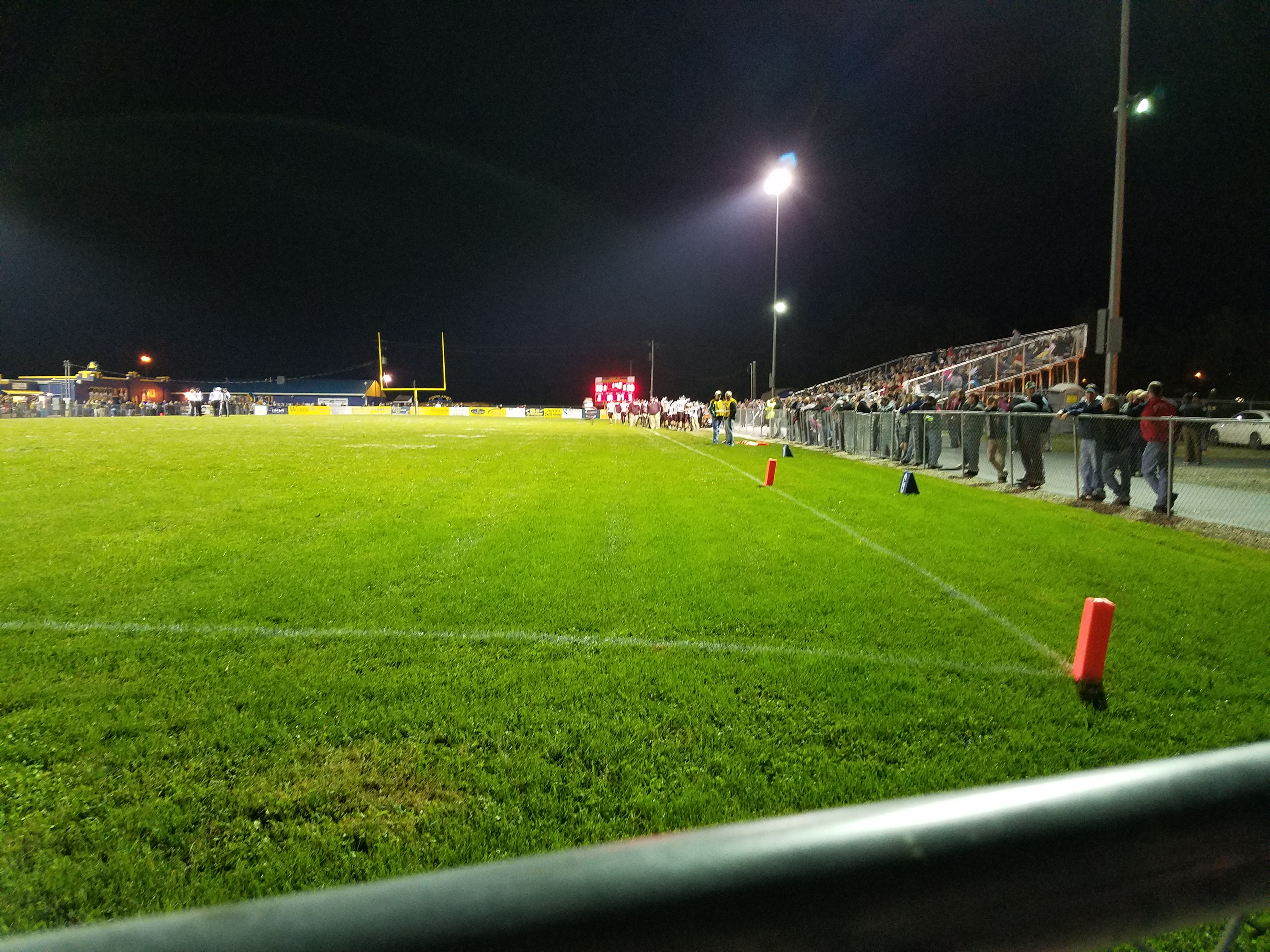 Night high school football game viewed from field level with stadium lights and sideline spectators.
