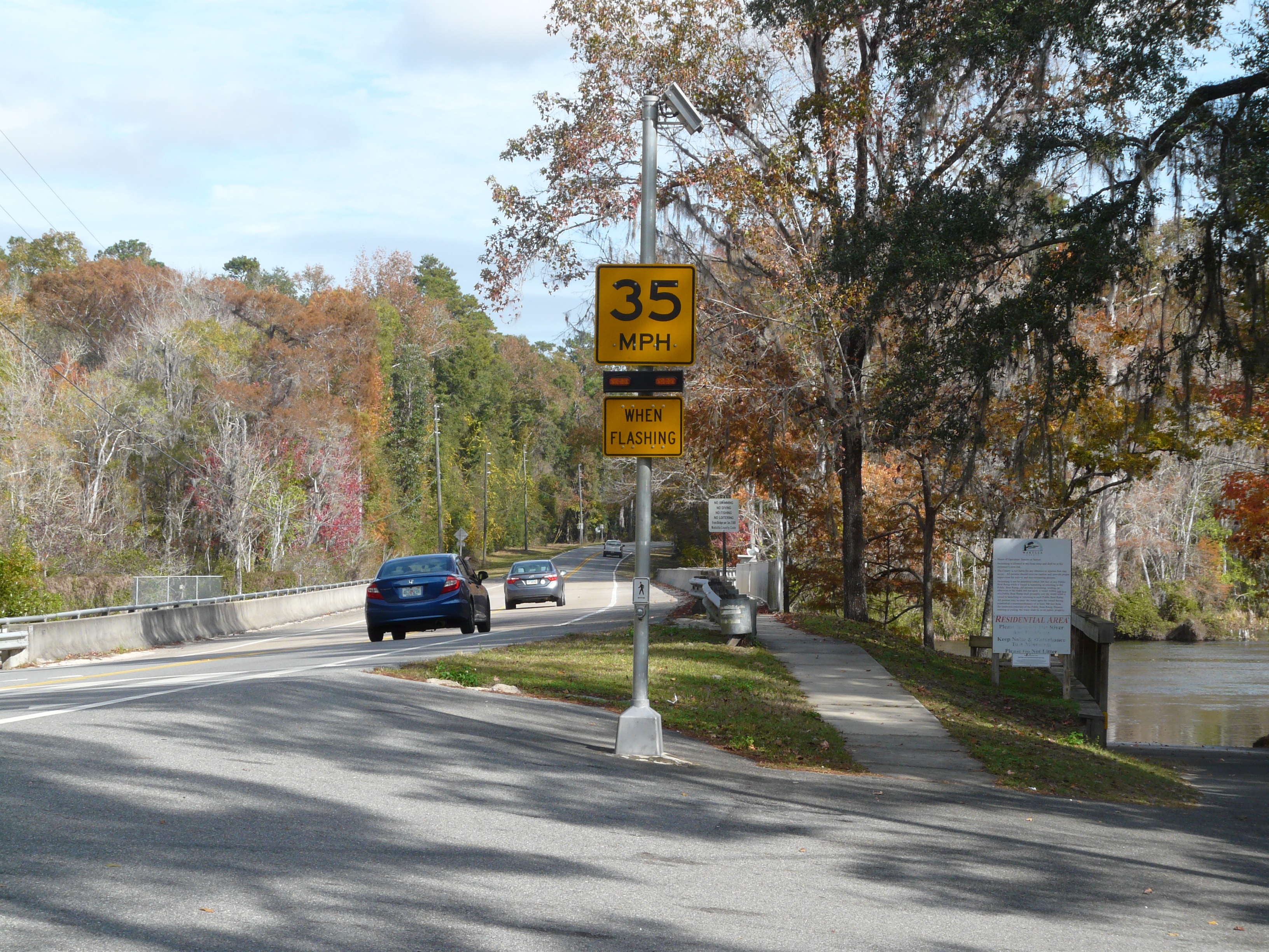 A rectangular rapid flashing beacon unit on a pole beside a two-lane road.