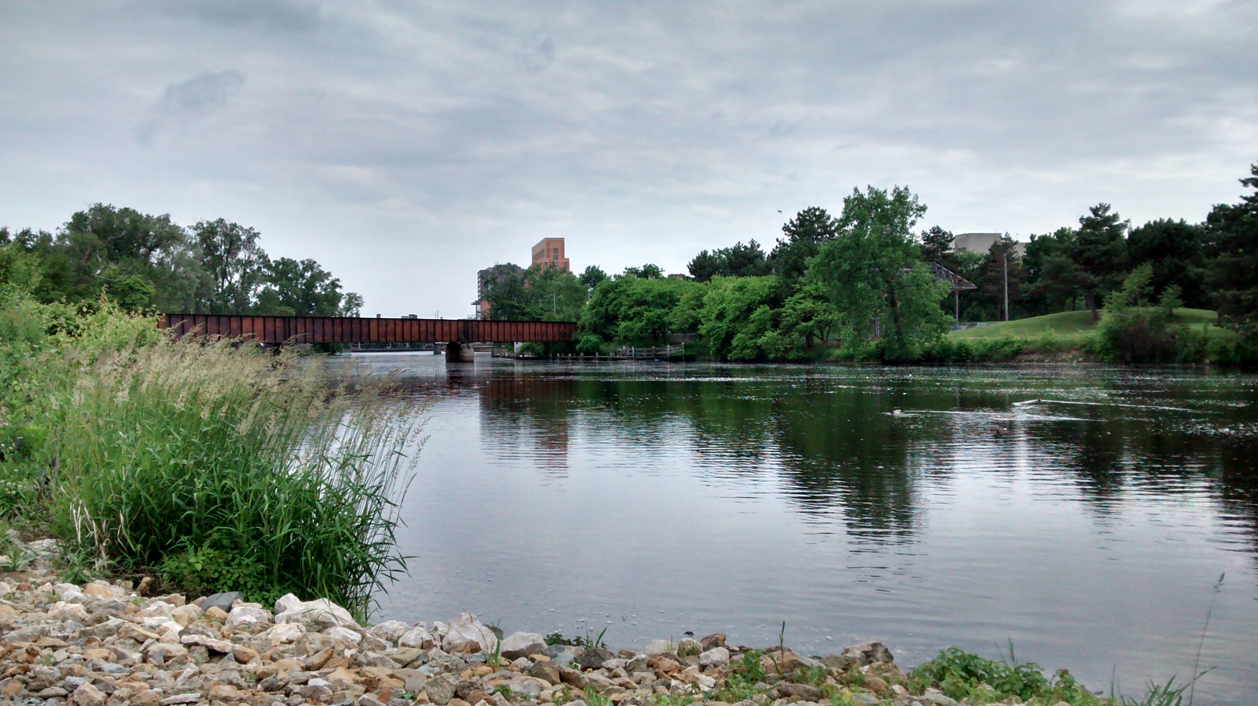 Metal truss bridge spanning a calm river with tree-lined banks and a rocky shoreline.