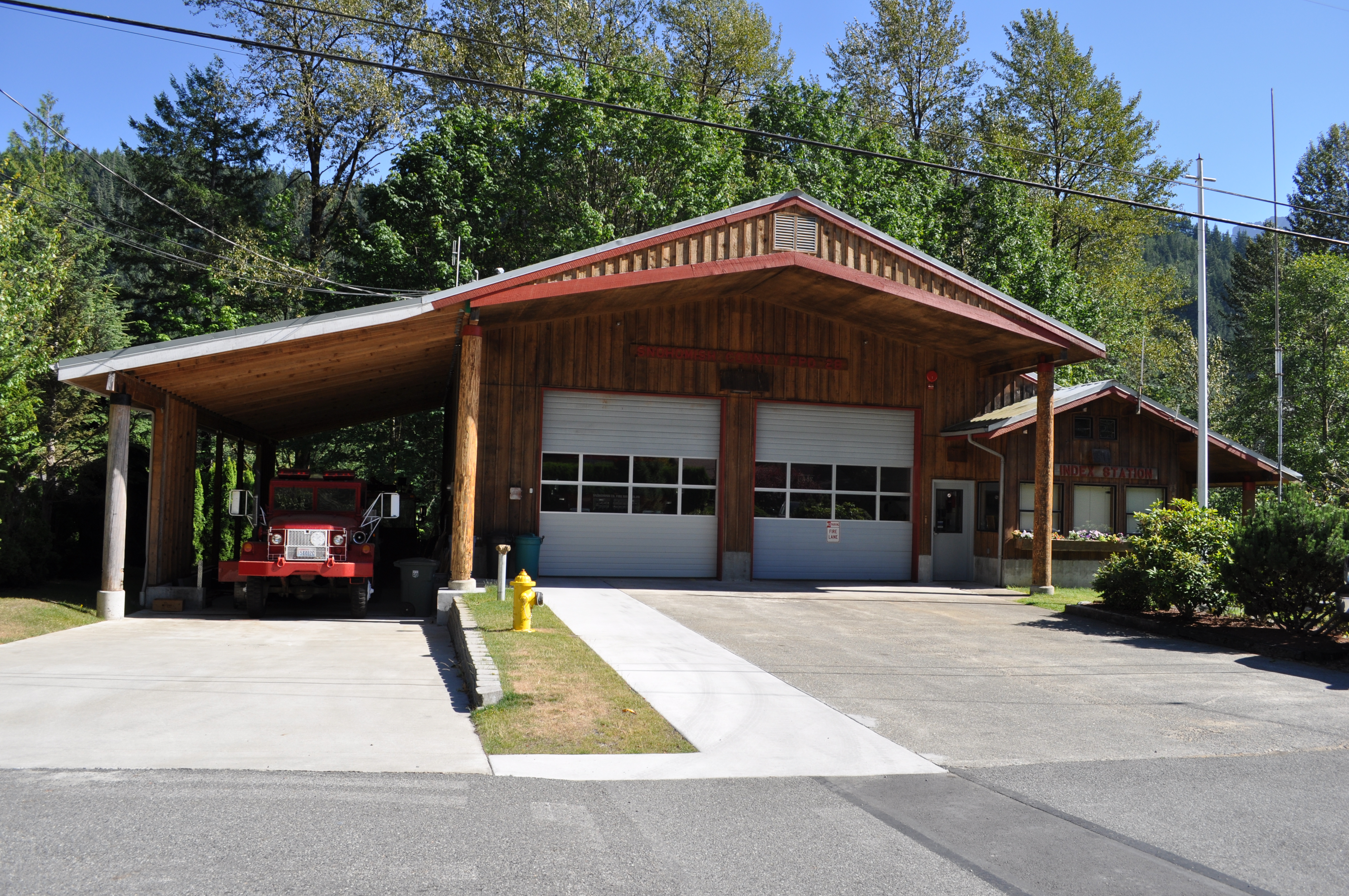 Wood-sided rural fire station with two garage bays and a red fire vehicle, surrounded by evergreen trees and hills.