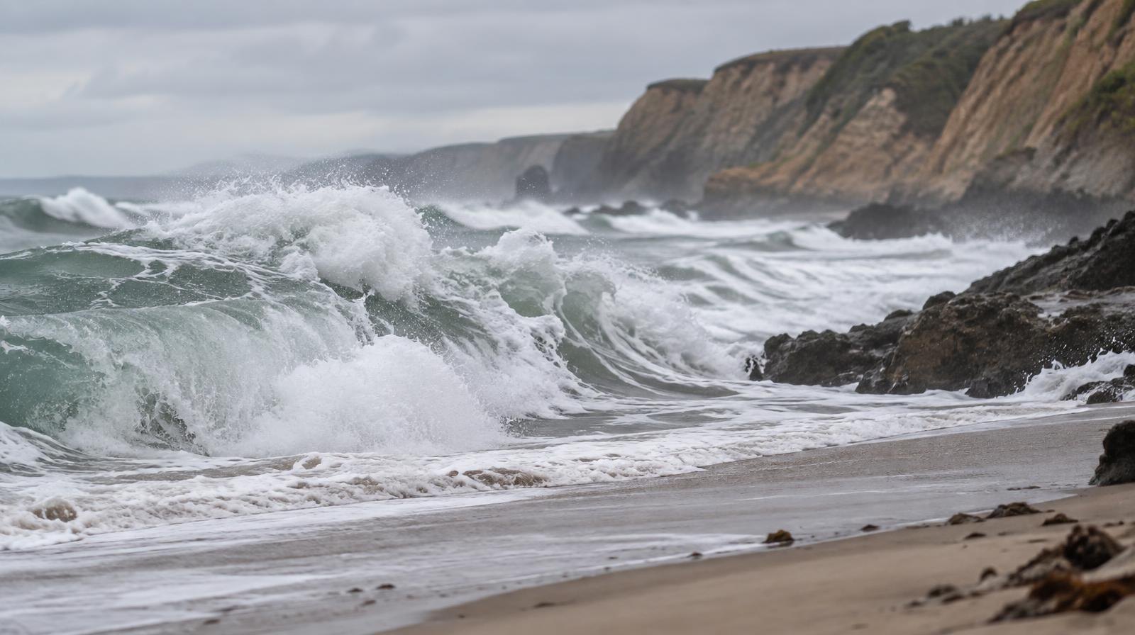 Large waves crash onto a sandy beach with eroded bluffs visible in the background.