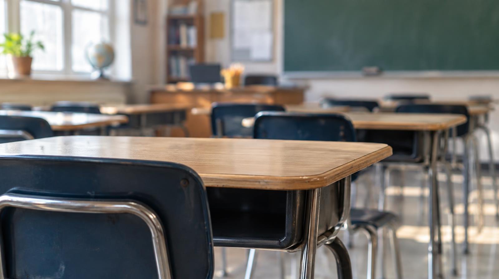 Rows of empty student desks in a sunlit classroom with a chalkboard at the front.