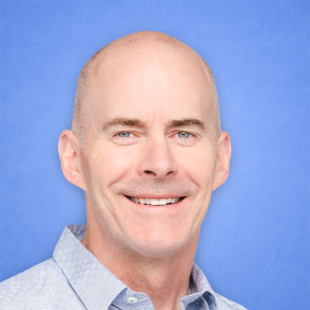 Professional headshot of a bald man in a light blue shirt smiling against a blue background.