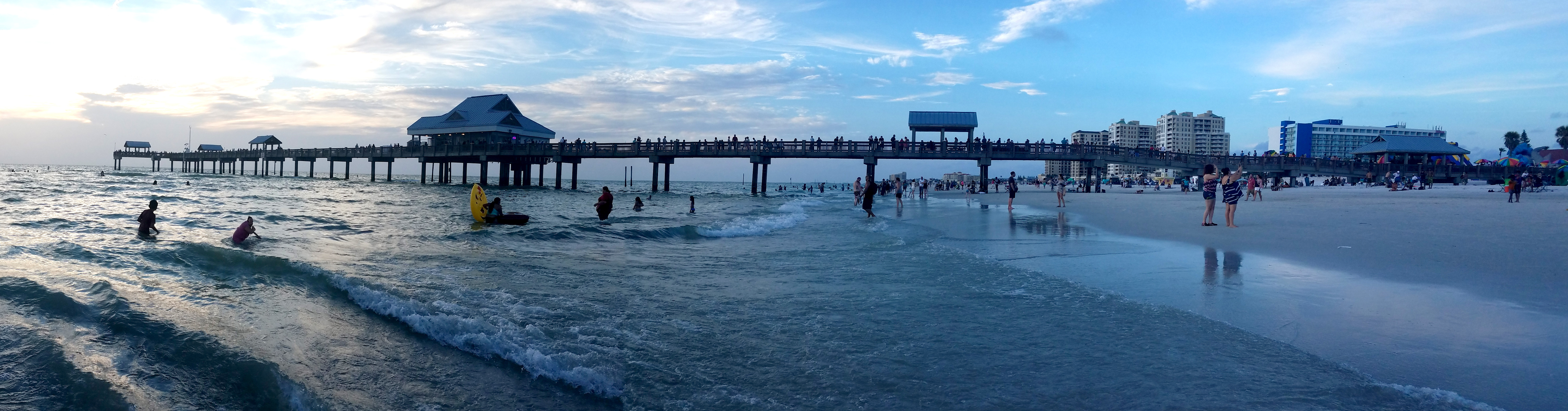Panoramic view of a pier stretching into calm Gulf waters at a busy Florida beach.