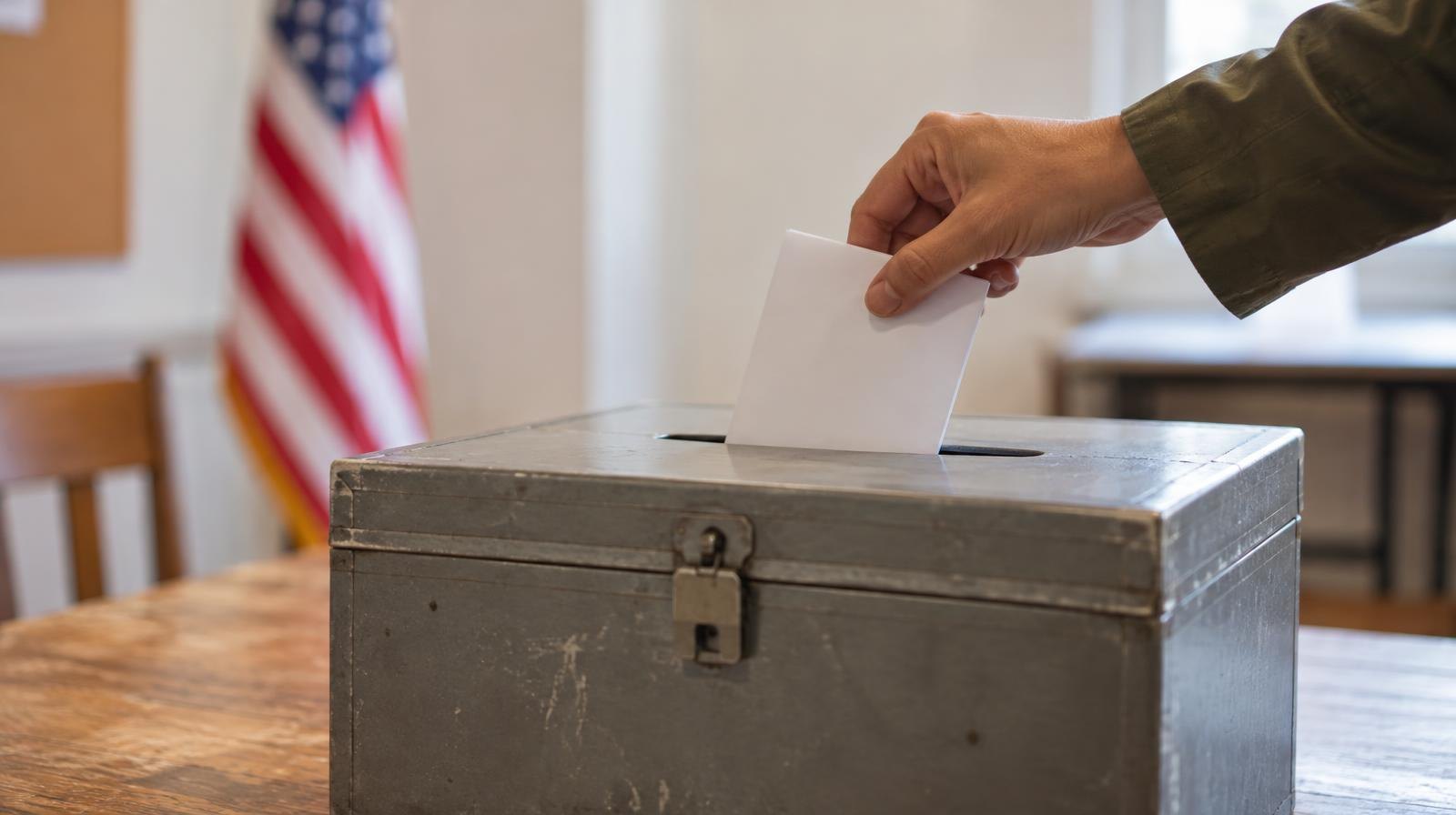 A hand placing a folded paper ballot into a metal ballot box on a wooden table.
